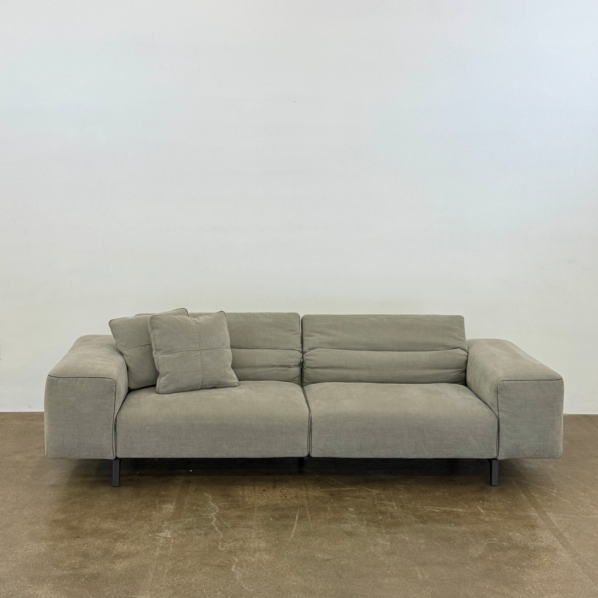 A modern Cassina Scighera Sofa by Cassina, in light gray with two matching square cushions, is placed on a bare concrete floor against a plain white wall.