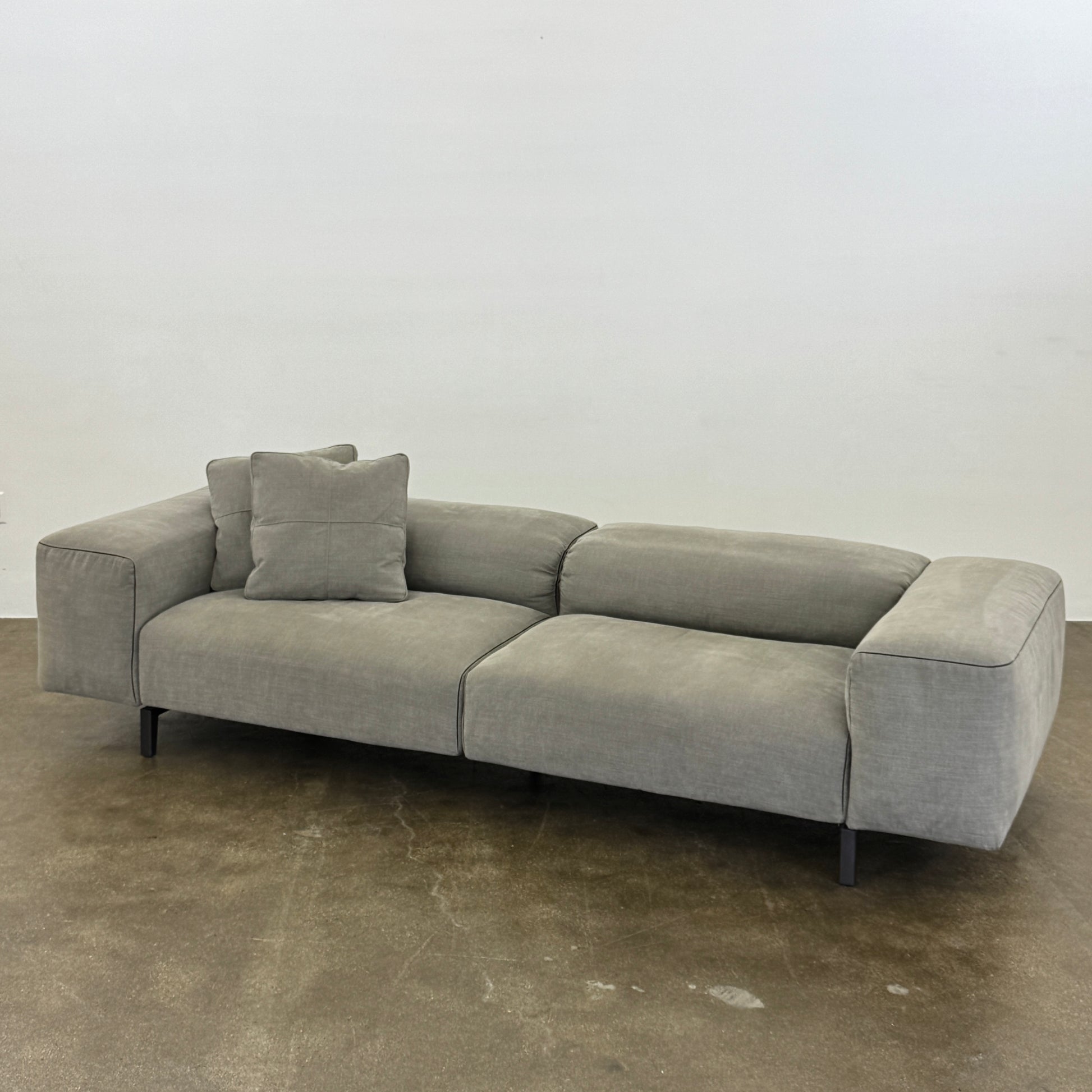 A modern Cassina Scighera Sofa by Cassina, in light gray with two matching square cushions, is placed on a bare concrete floor against a plain white wall.