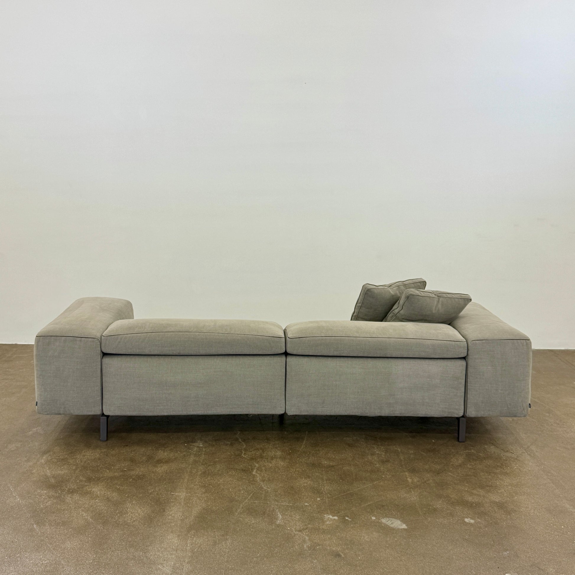 A modern Cassina Scighera Sofa by Cassina, in light gray with two matching square cushions, is placed on a bare concrete floor against a plain white wall.