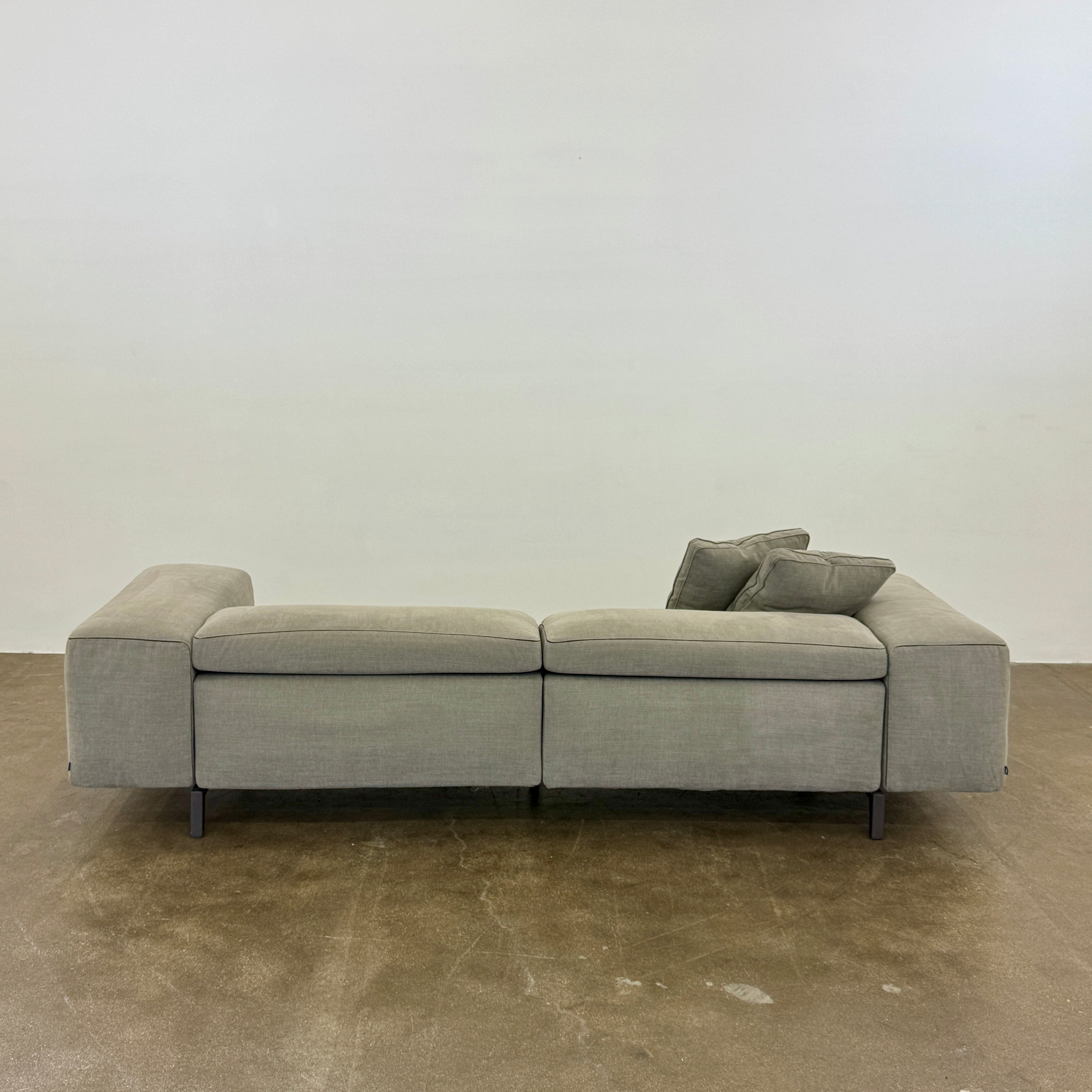 A modern Cassina Scighera Sofa by Cassina, in light gray with two matching square cushions, is placed on a bare concrete floor against a plain white wall.
