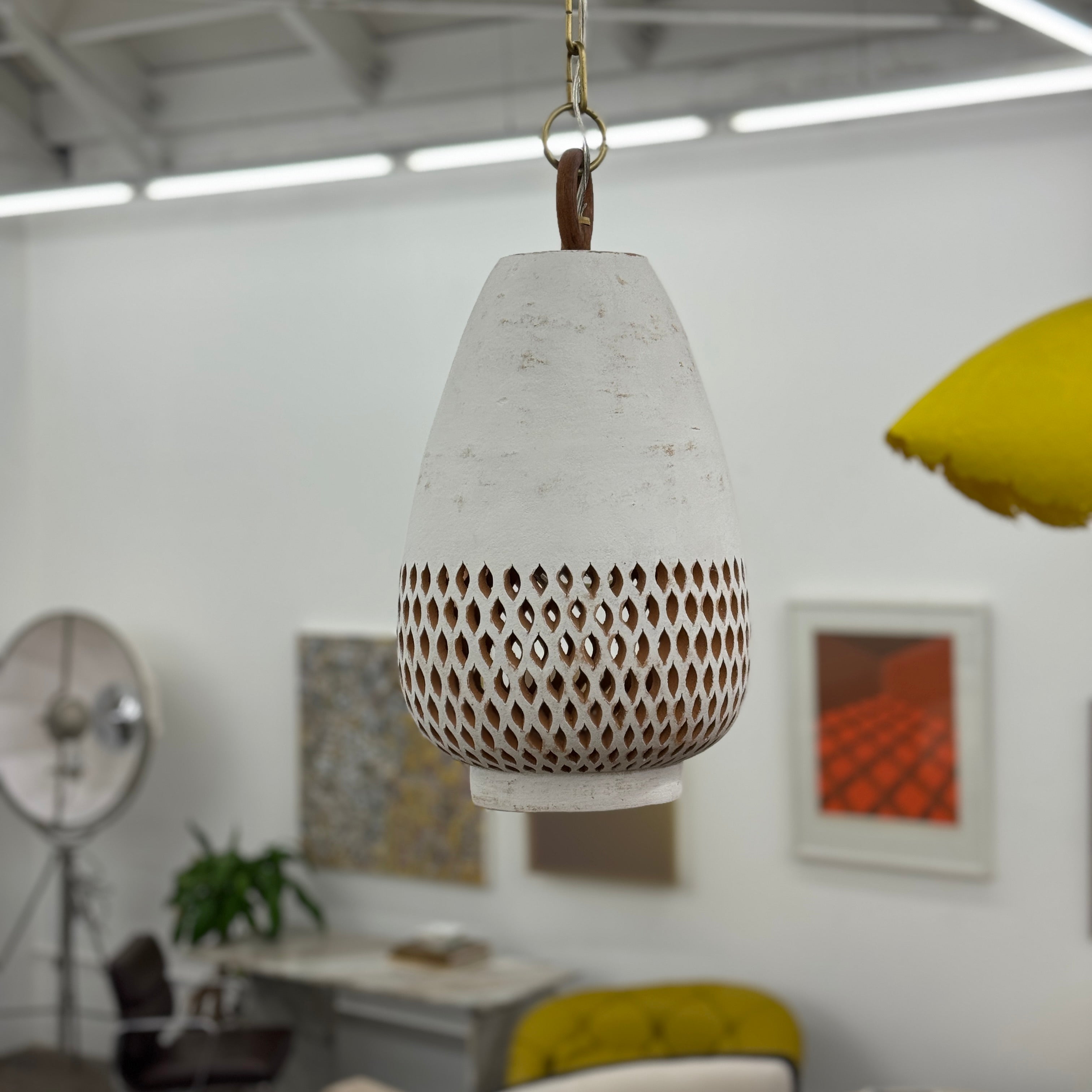The L’aviva Diamantes Pendant Light, Size B by L’aviva features a white ceramic shade with hand-cut clay patterns, inspired by Oaxacan pottery, and hangs in a modern room with art and contemporary furniture.