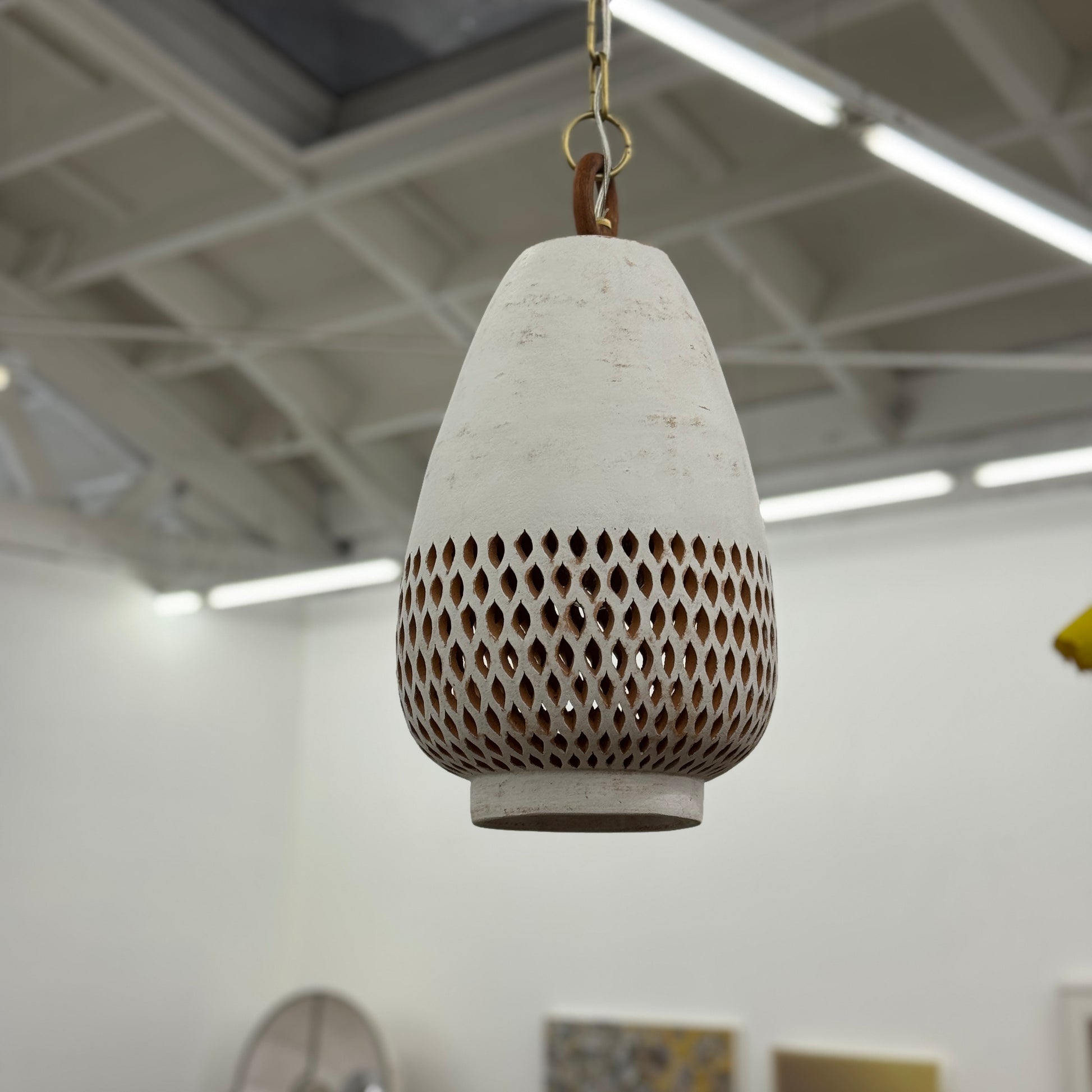 The L’aviva Diamantes Pendant Light, Size B by L’aviva features a white ceramic shade with hand-cut clay patterns, inspired by Oaxacan pottery, and hangs in a modern room with art and contemporary furniture.