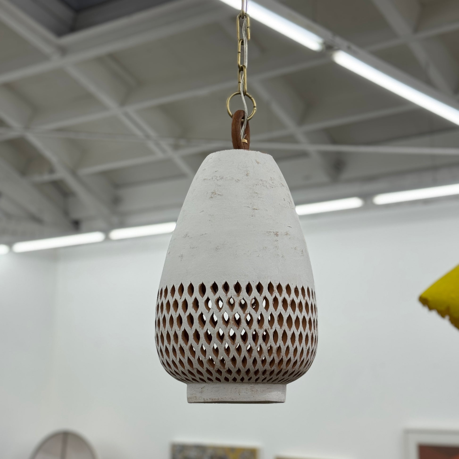 The L’aviva Diamantes Pendant Light, Size B by L’aviva features a white ceramic shade with hand-cut clay patterns, inspired by Oaxacan pottery, and hangs in a modern room with art and contemporary furniture.