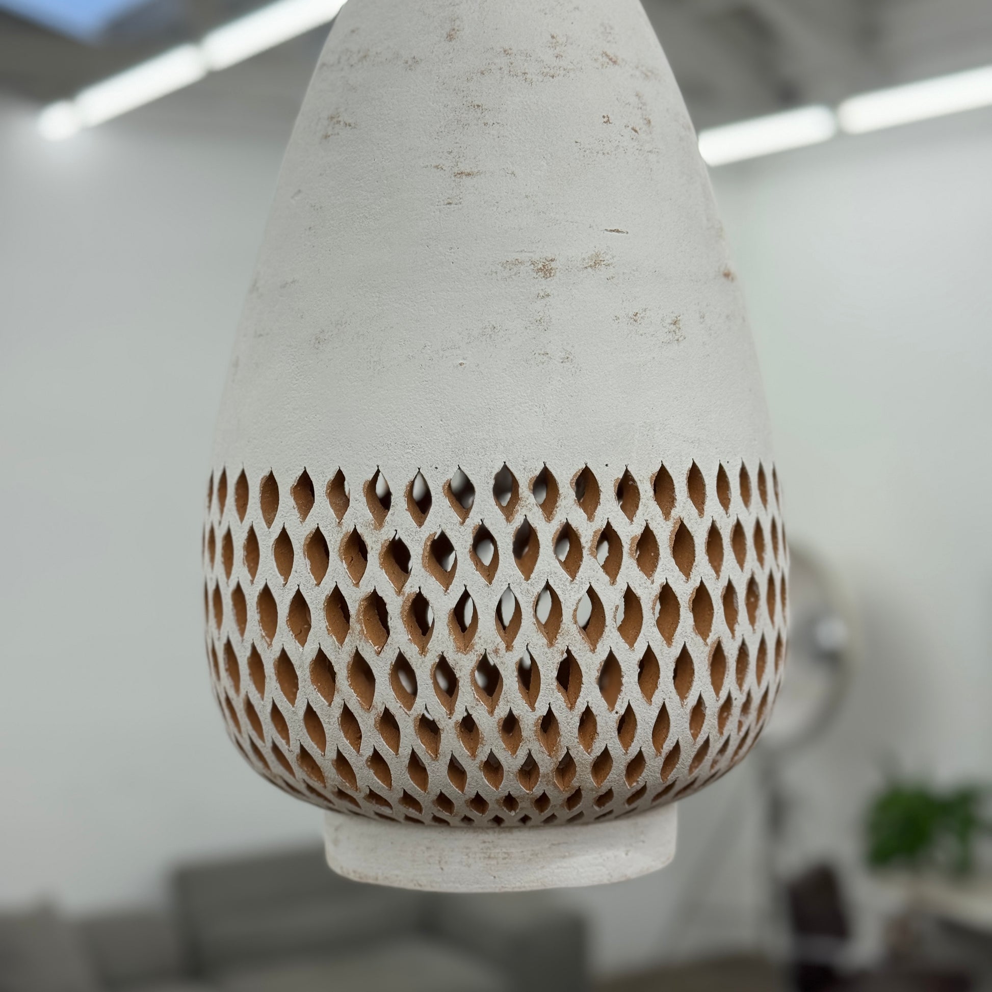 The L’aviva Diamantes Pendant Light, Size B by L’aviva features a white ceramic shade with hand-cut clay patterns, inspired by Oaxacan pottery, and hangs in a modern room with art and contemporary furniture.