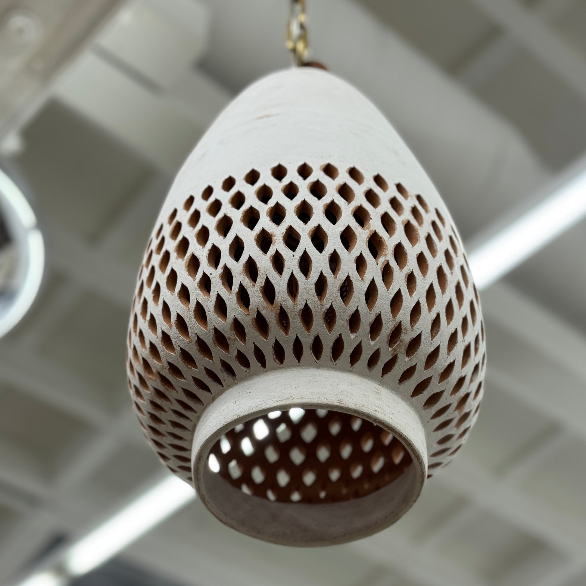 The L’aviva Diamantes Pendant Light, Size B by L’aviva features a white ceramic shade with hand-cut clay patterns, inspired by Oaxacan pottery, and hangs in a modern room with art and contemporary furniture.