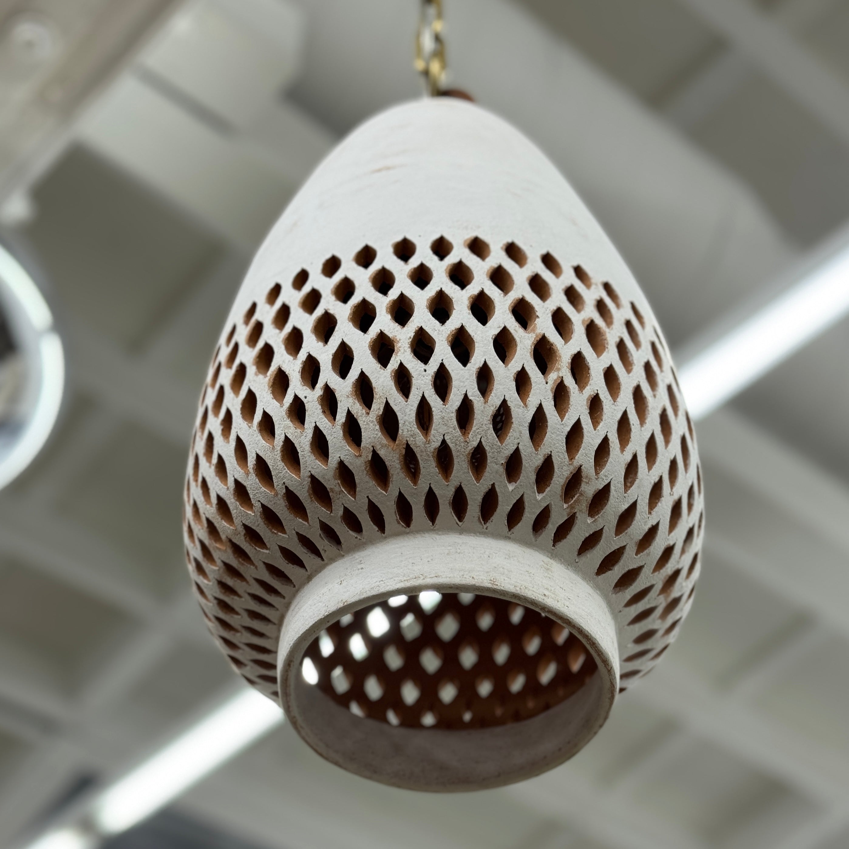 The L’aviva Diamantes Pendant Light, Size B by L’aviva features a white ceramic shade with hand-cut clay patterns, inspired by Oaxacan pottery, and hangs in a modern room with art and contemporary furniture.