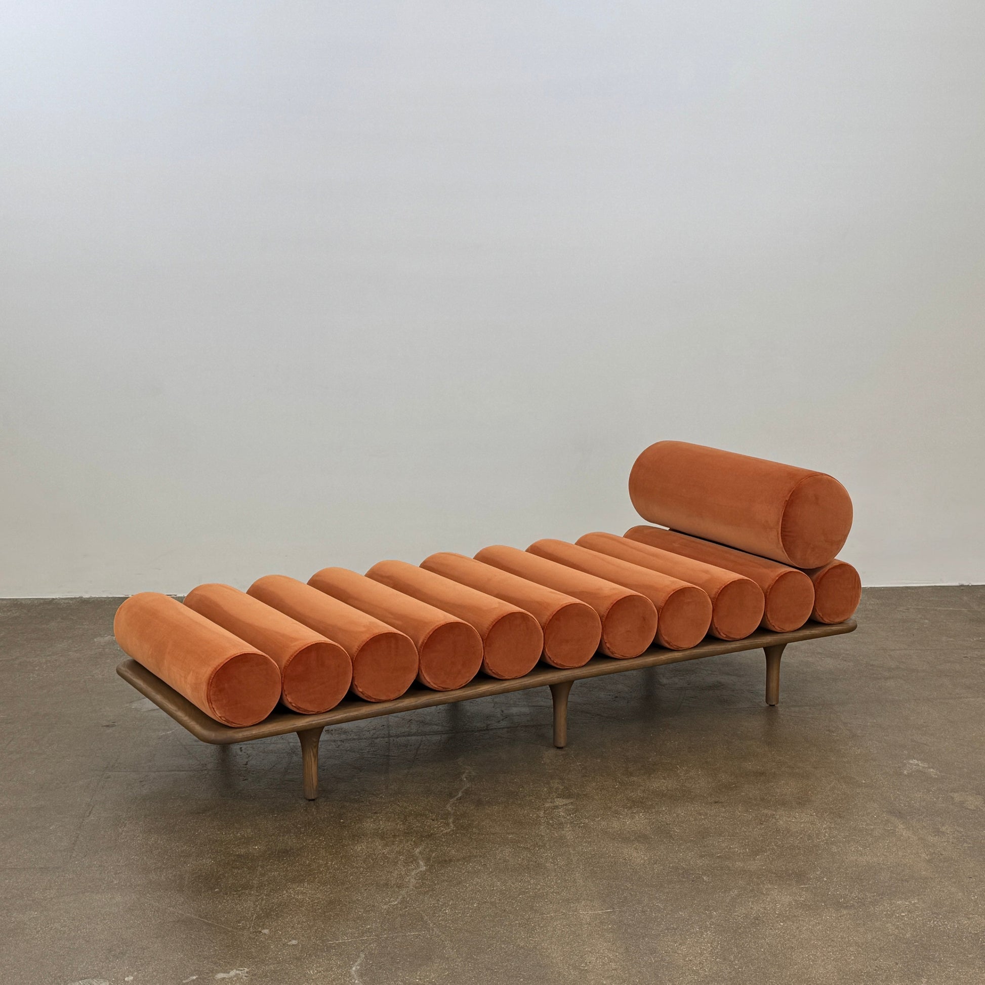 The Tacchini Five to Nine Day Bed by Tacchini features orange cylindrical cushions on a wooden frame—an ideal modern piece for an artist studio, shown here against a plain white background.