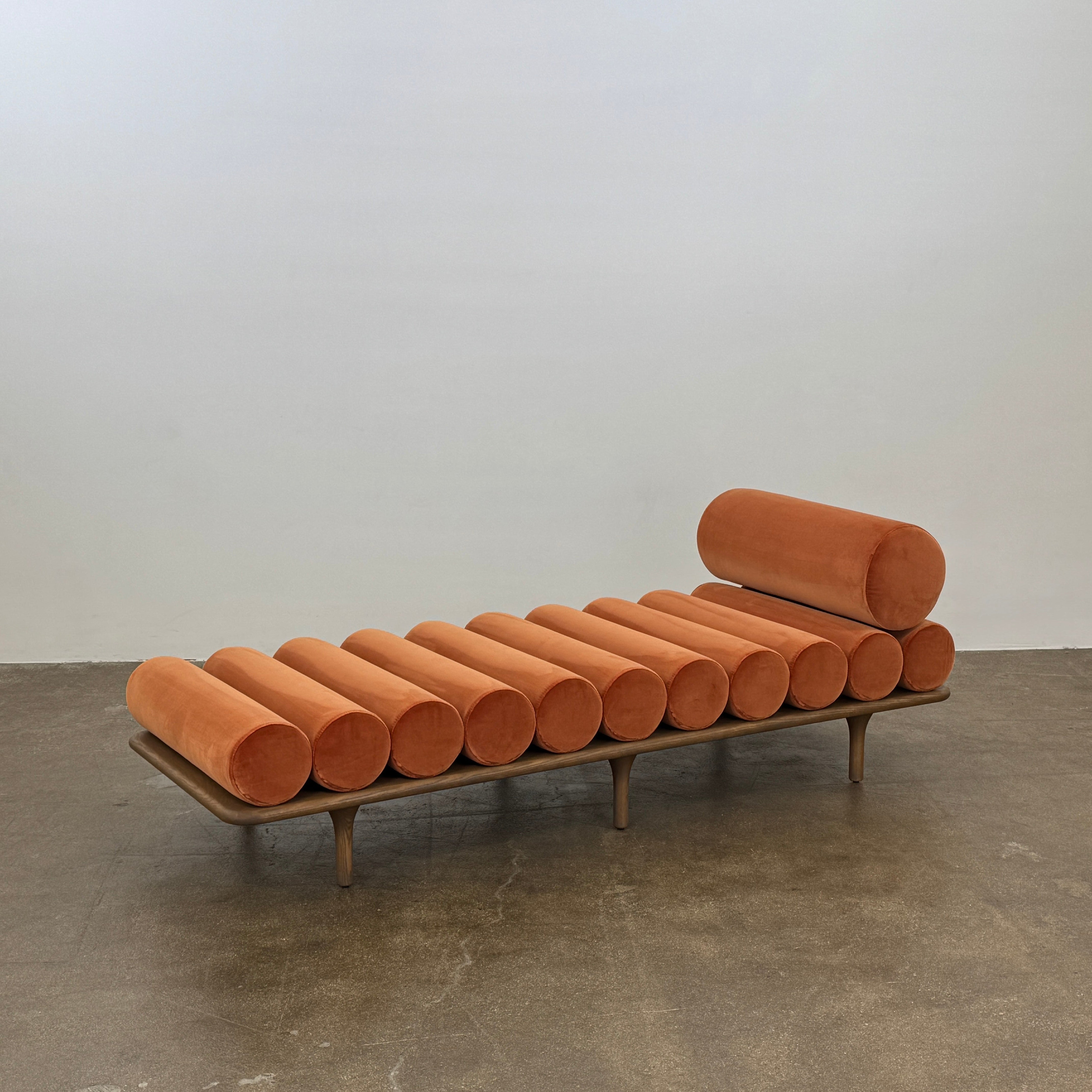 The Tacchini Five to Nine Day Bed by Tacchini features orange cylindrical cushions on a wooden frame—an ideal modern piece for an artist studio, shown here against a plain white background.