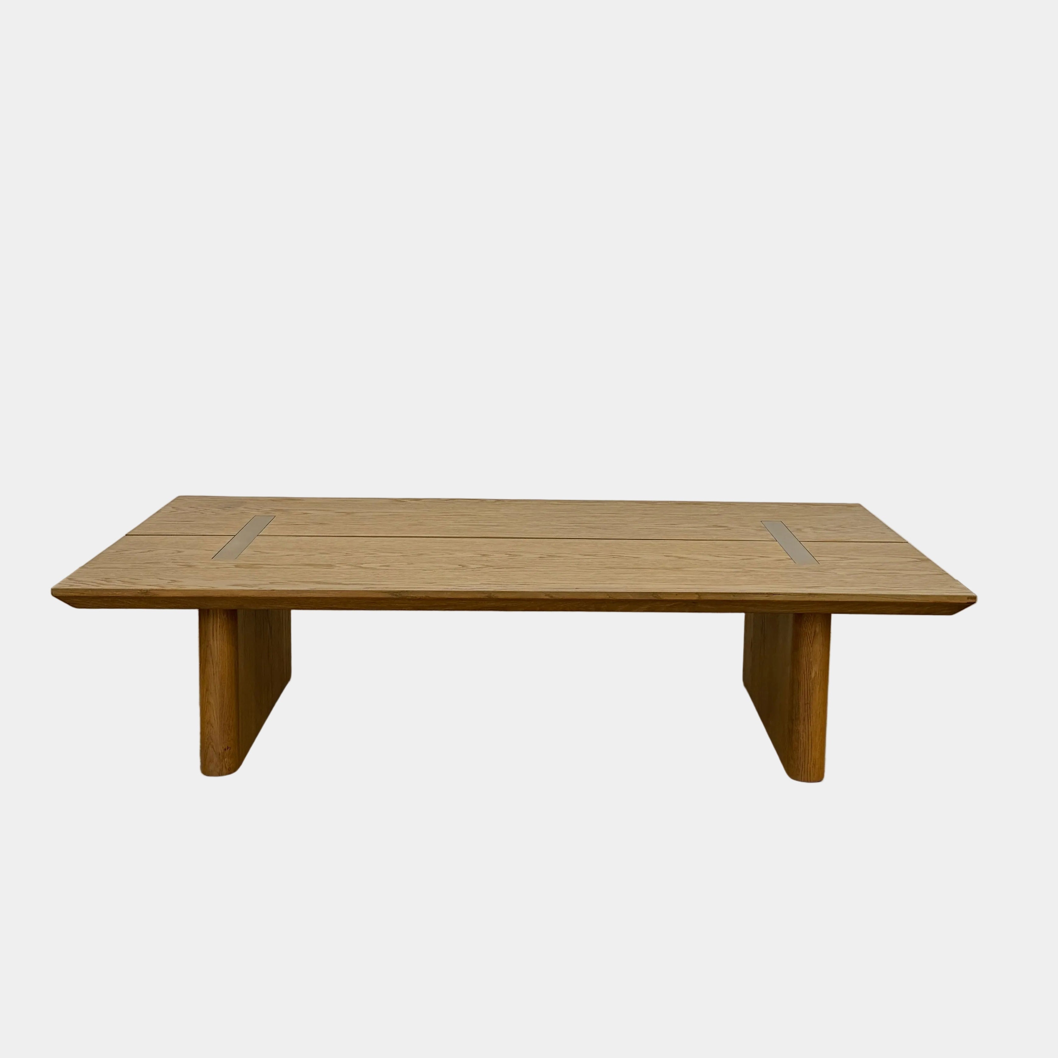 The Harbour Rozzell Coffee Table by Harbour features a rectangular wooden design with two wide legs, set on a concrete floor beside a plain white wall.