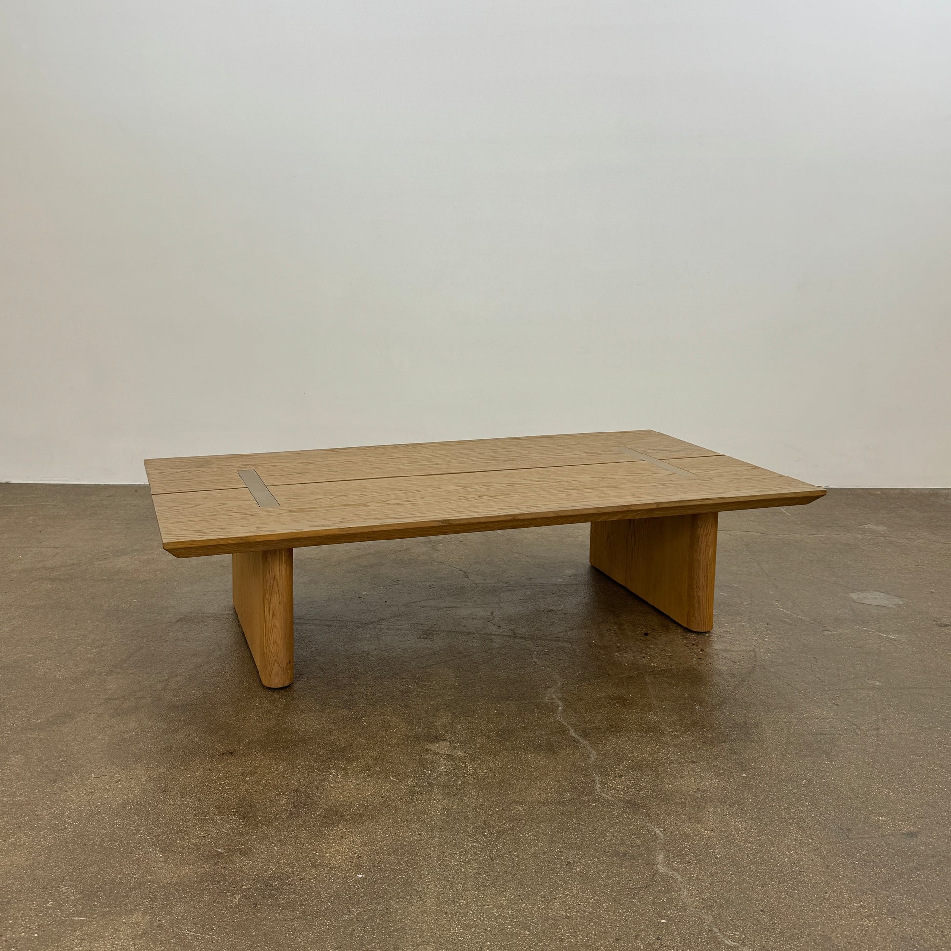 The Harbour Rozzell Coffee Table by Harbour features a rectangular wooden design with two wide legs, set on a concrete floor beside a plain white wall.