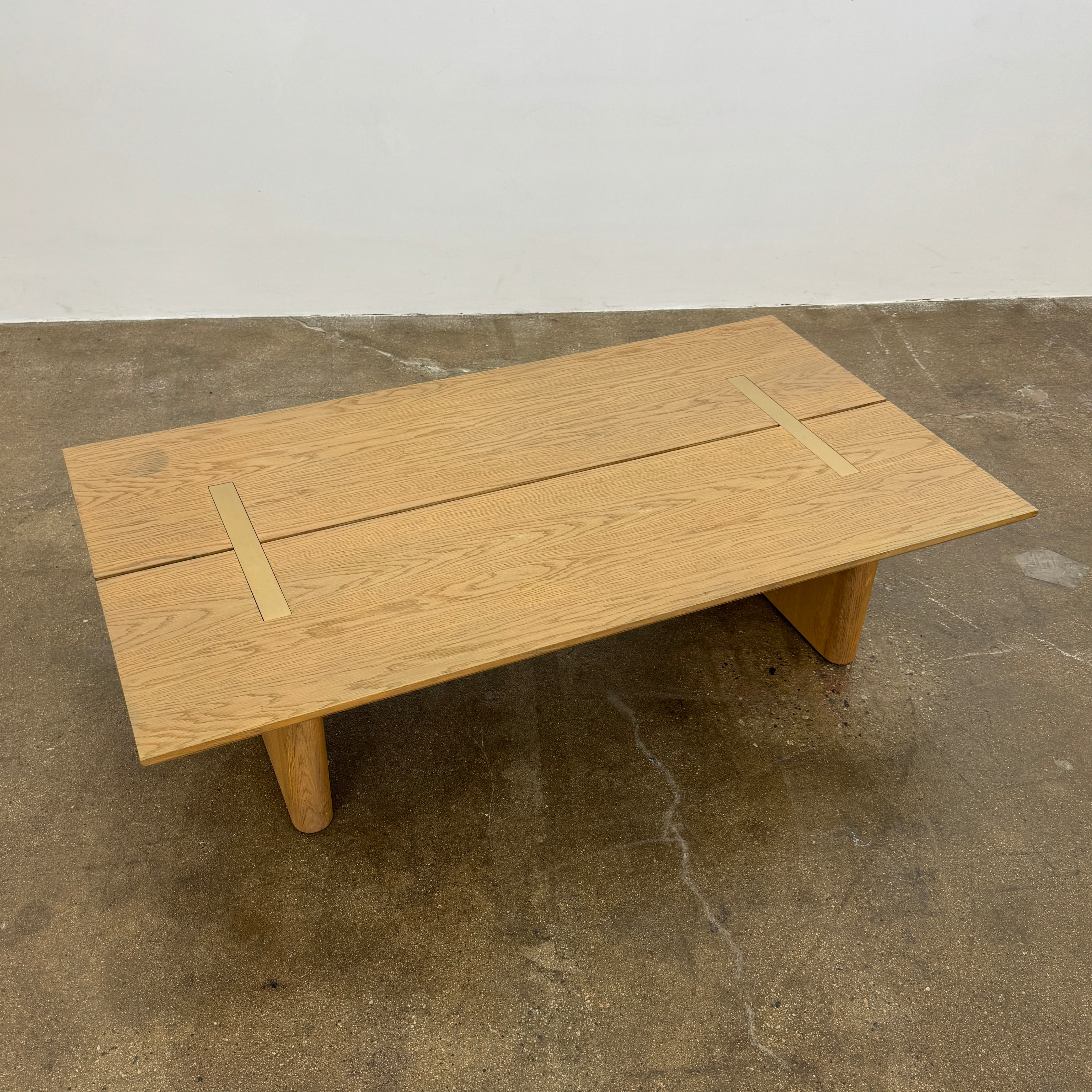 The Harbour Rozzell Coffee Table by Harbour features a rectangular wooden design with two wide legs, set on a concrete floor beside a plain white wall.