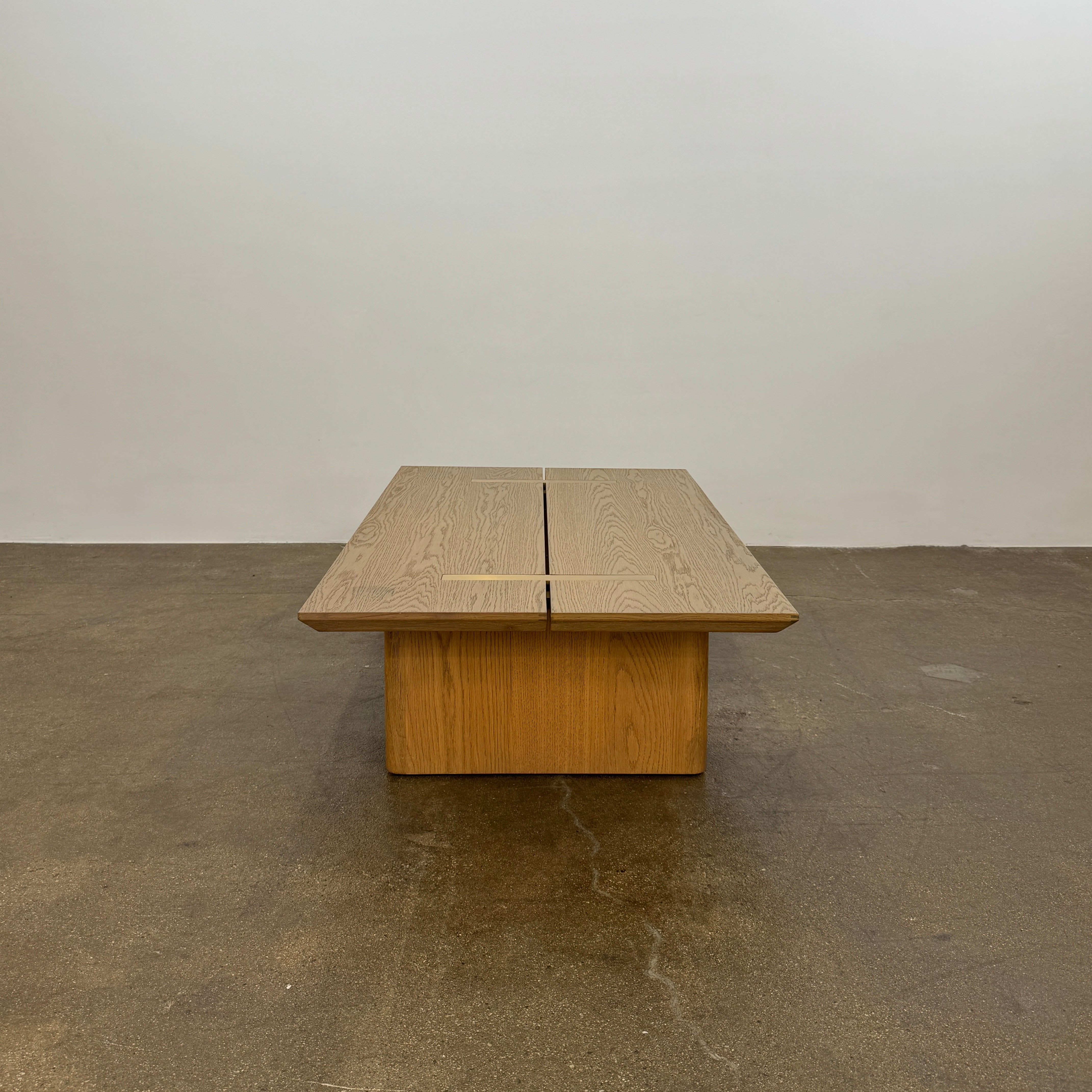 The Harbour Rozzell Coffee Table by Harbour features a rectangular wooden design with two wide legs, set on a concrete floor beside a plain white wall.