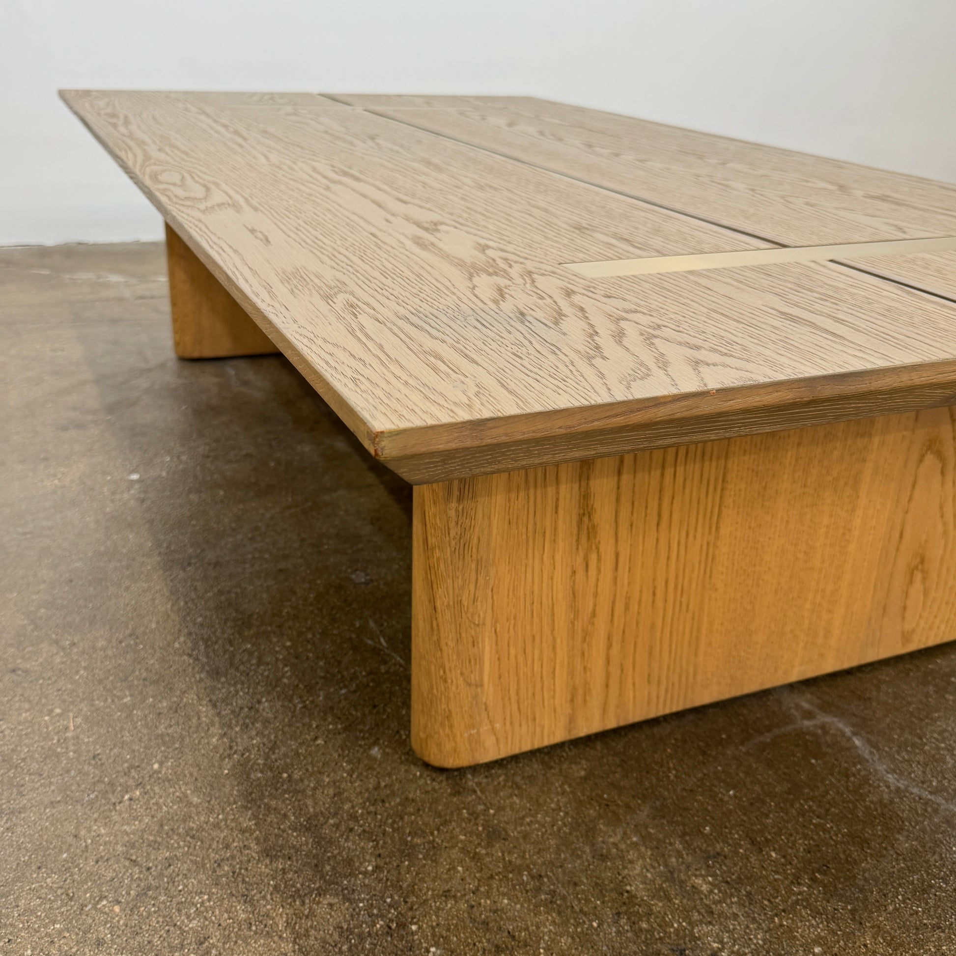 The Harbour Rozzell Coffee Table by Harbour features a rectangular wooden design with two wide legs, set on a concrete floor beside a plain white wall.