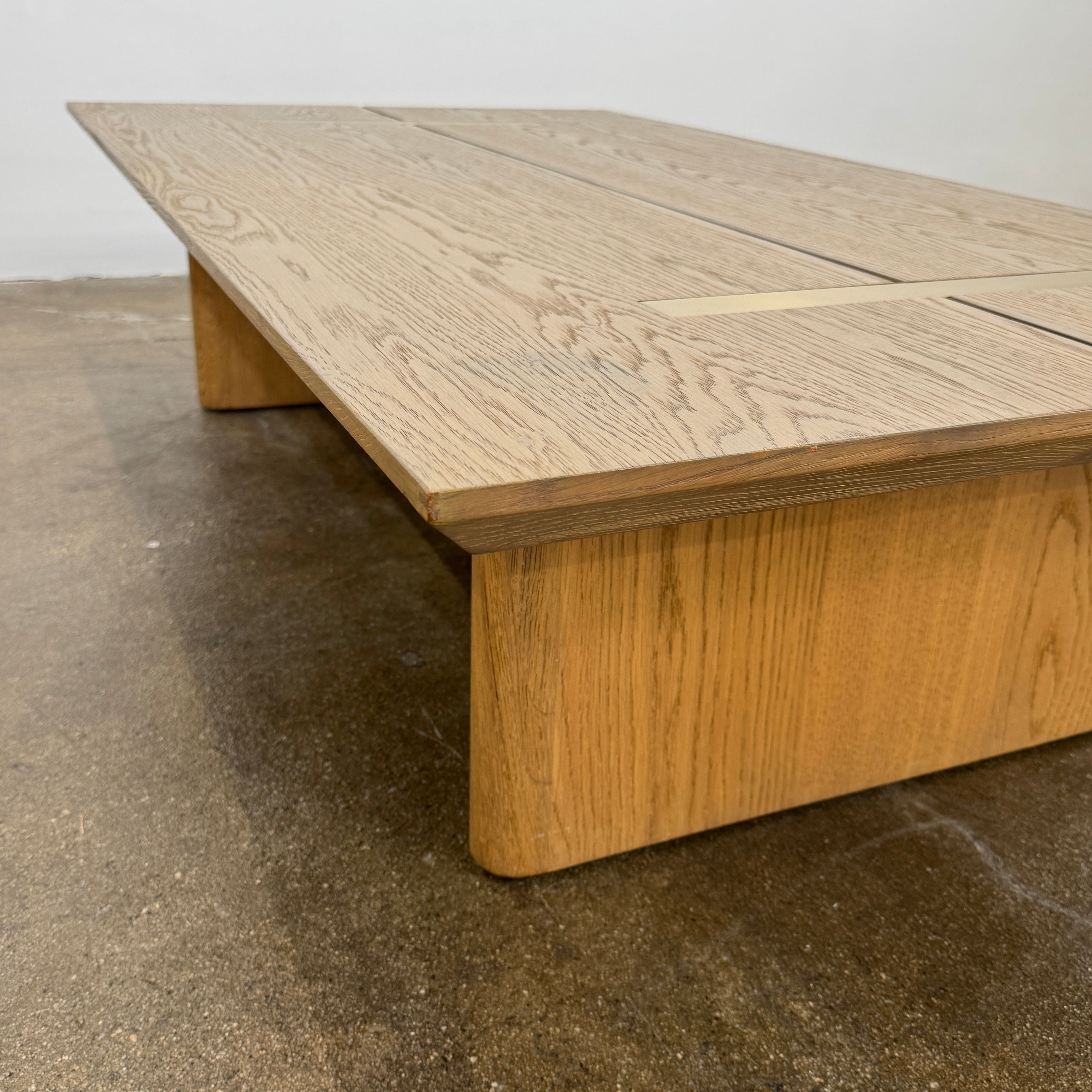 The Harbour Rozzell Coffee Table by Harbour features a rectangular wooden design with two wide legs, set on a concrete floor beside a plain white wall.