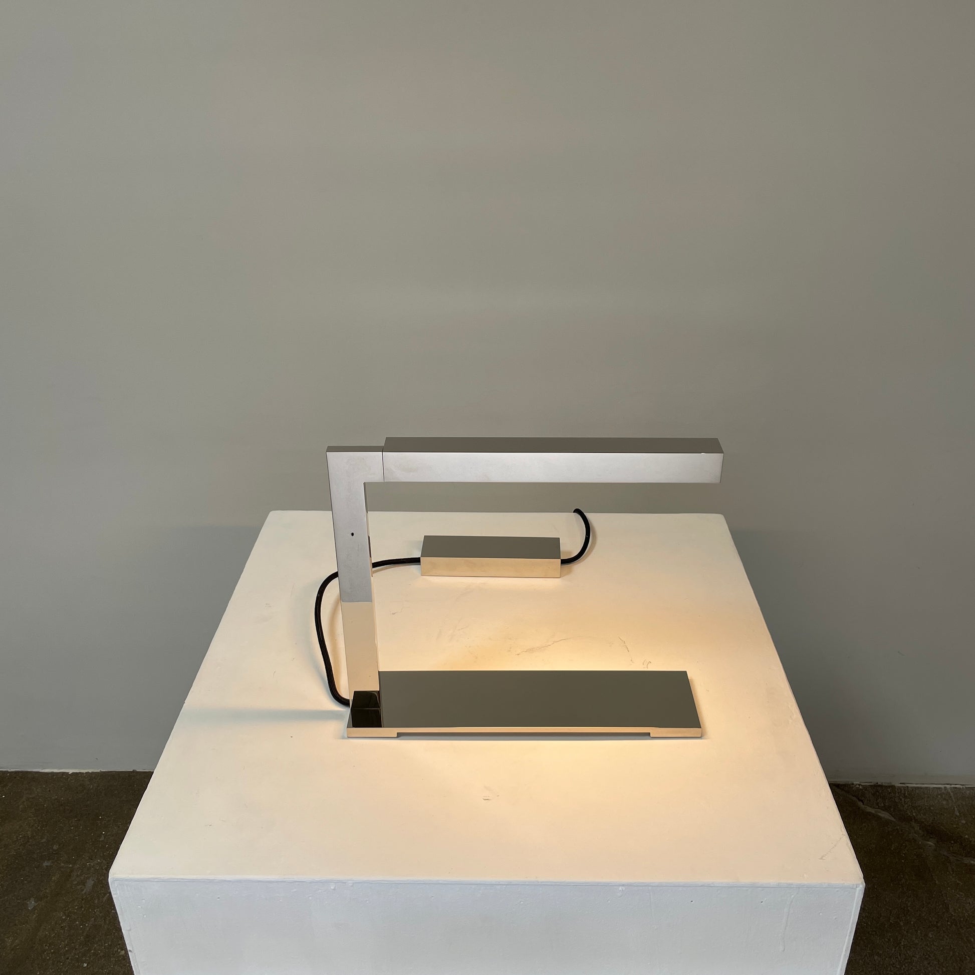 The Holly Hunt Studio Bar Desk Lamp, featuring a modern minimalist design with a rectangular base and adjustable arm, is elegantly placed on a white pedestal against a gray wall.