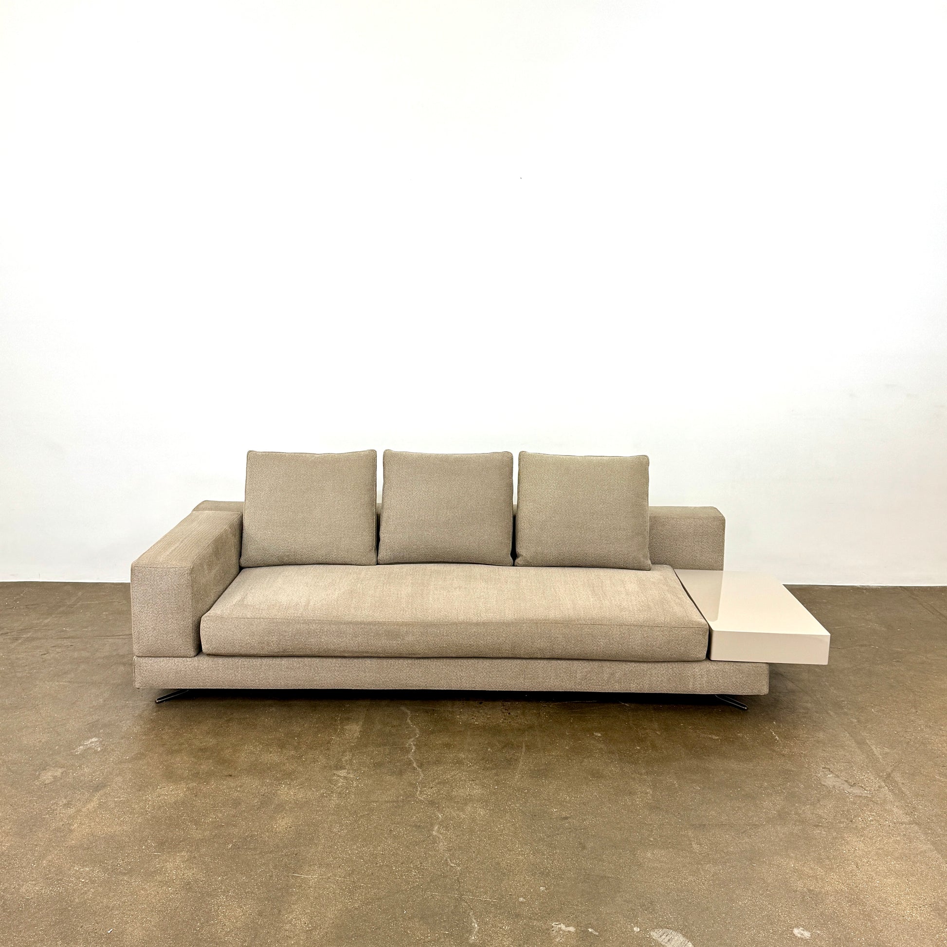 The Minotti White Sofa with Table is a beige three-seater luxury sofa featuring chenille upholstery, wide armrests, three back cushions, and an attached white side table on the right, set against a plain white wall on a brown floor.