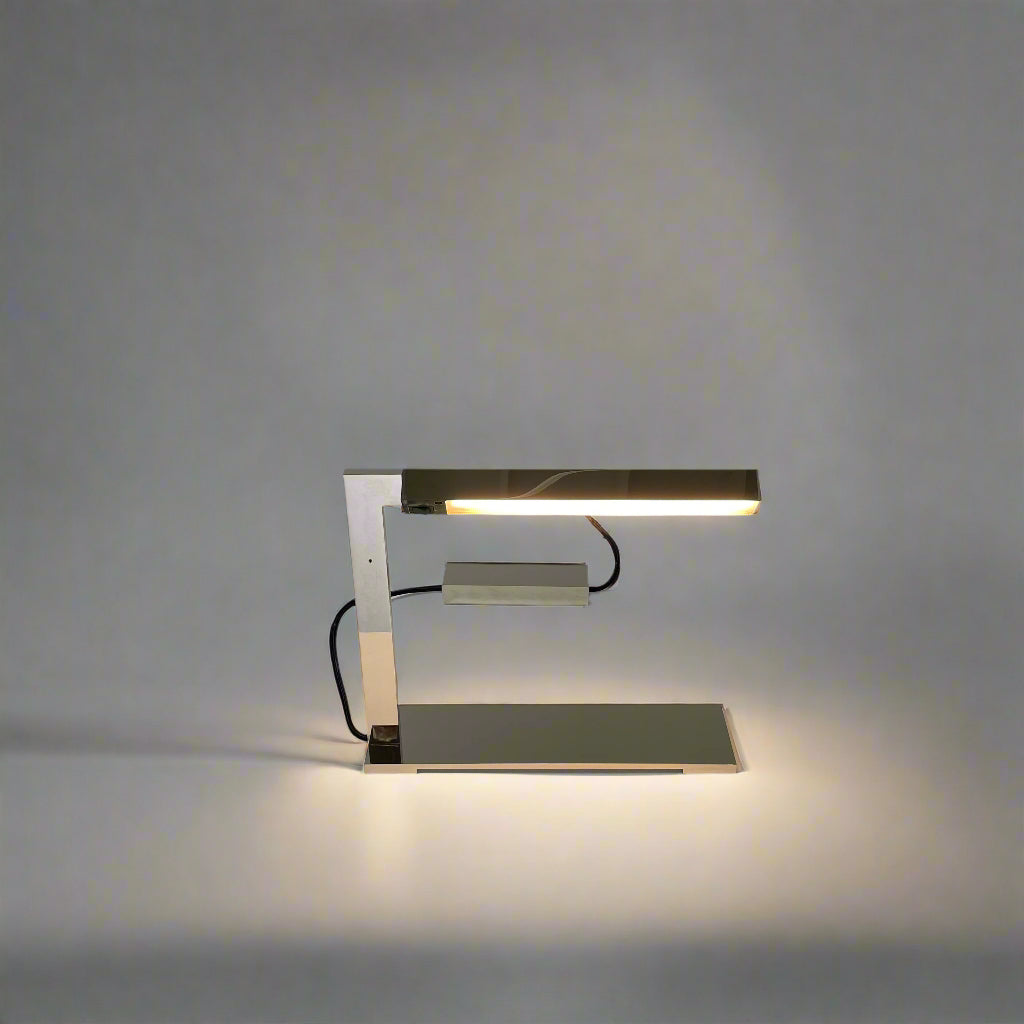 The Holly Hunt Studio Bar Desk Lamp, featuring a modern minimalist design with a rectangular base and adjustable arm, is elegantly placed on a white pedestal against a gray wall.