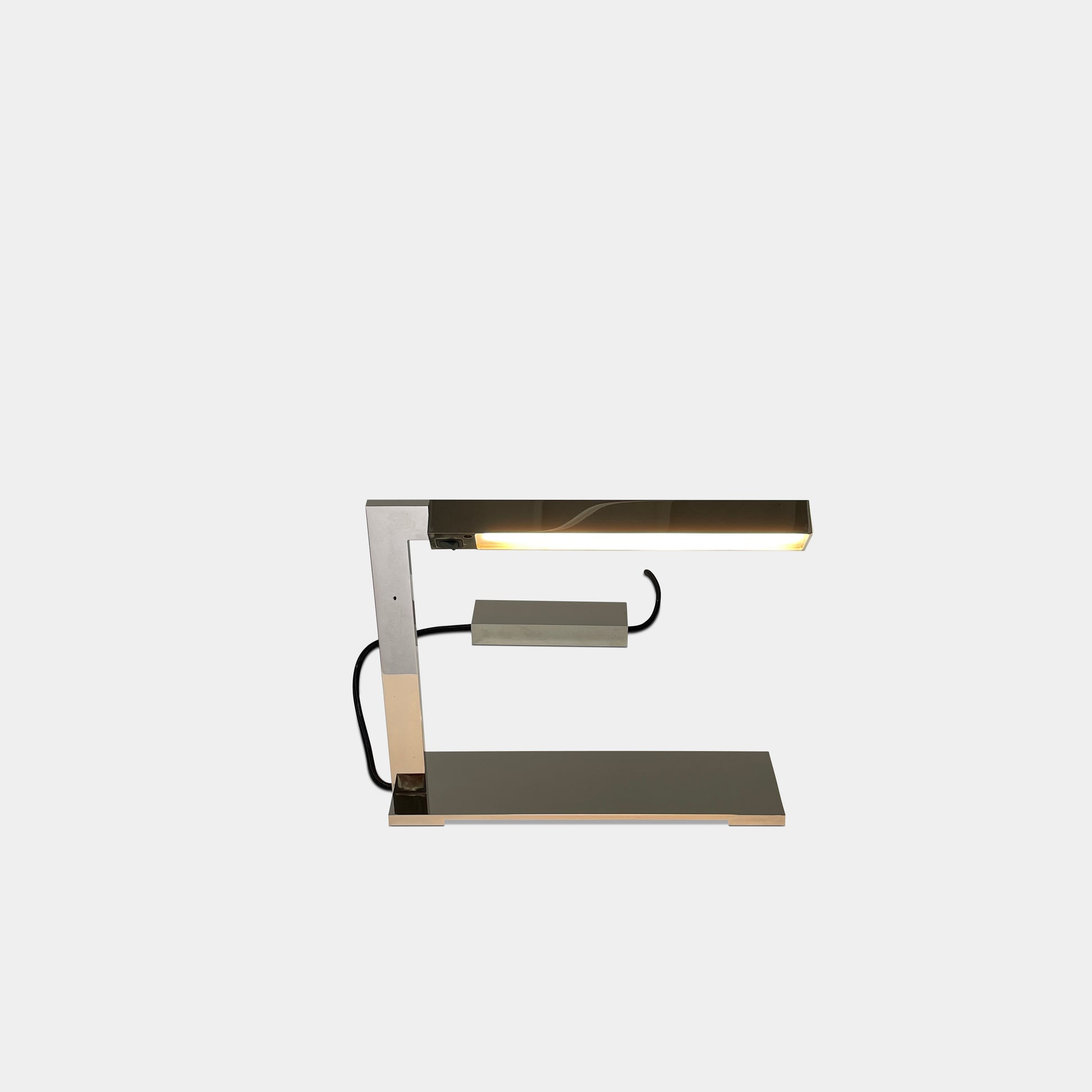 The Holly Hunt Studio Bar Desk Lamp, featuring a modern minimalist design with a rectangular base and adjustable arm, is elegantly placed on a white pedestal against a gray wall.