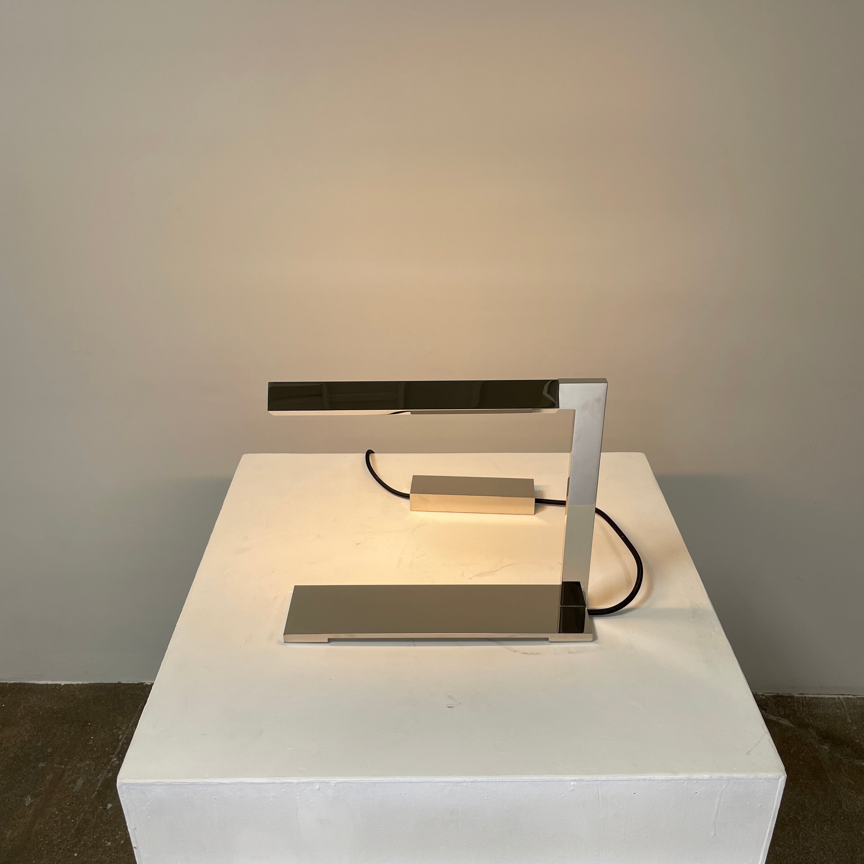 The Holly Hunt Studio Bar Desk Lamp, featuring a modern minimalist design with a rectangular base and adjustable arm, is elegantly placed on a white pedestal against a gray wall.