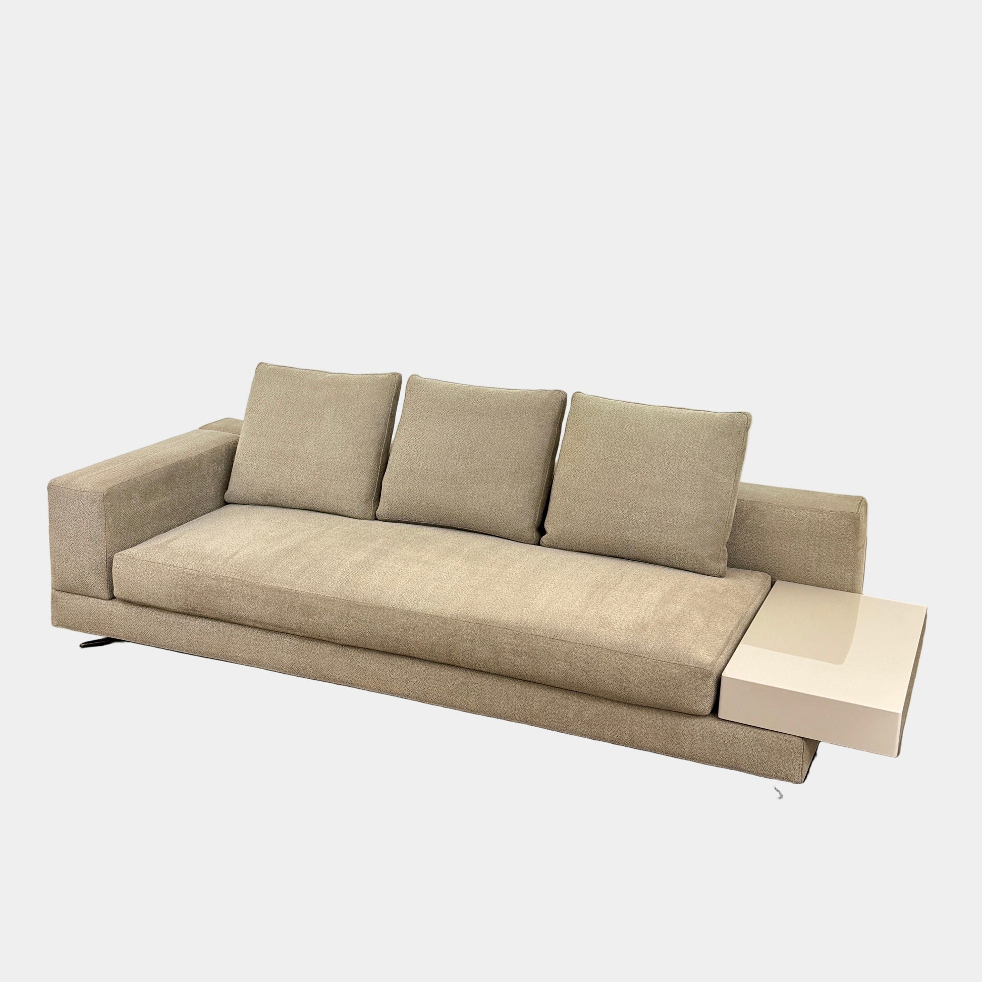 The Minotti White Sofa with Table is a beige three-seater luxury sofa featuring chenille upholstery, wide armrests, three back cushions, and an attached white side table on the right, set against a plain white wall on a brown floor.