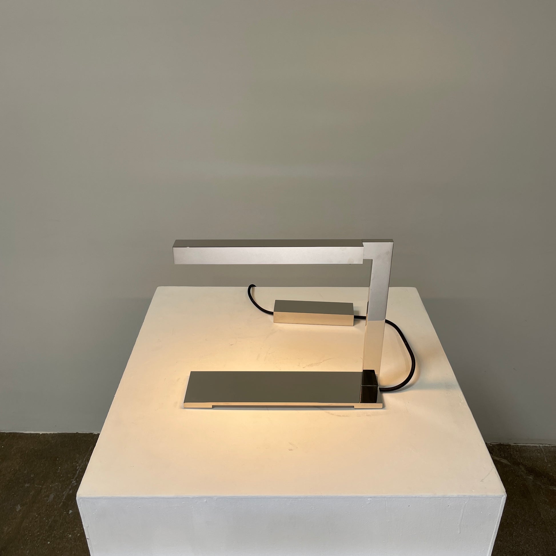 The Holly Hunt Studio Bar Desk Lamp, featuring a modern minimalist design with a rectangular base and adjustable arm, is elegantly placed on a white pedestal against a gray wall.