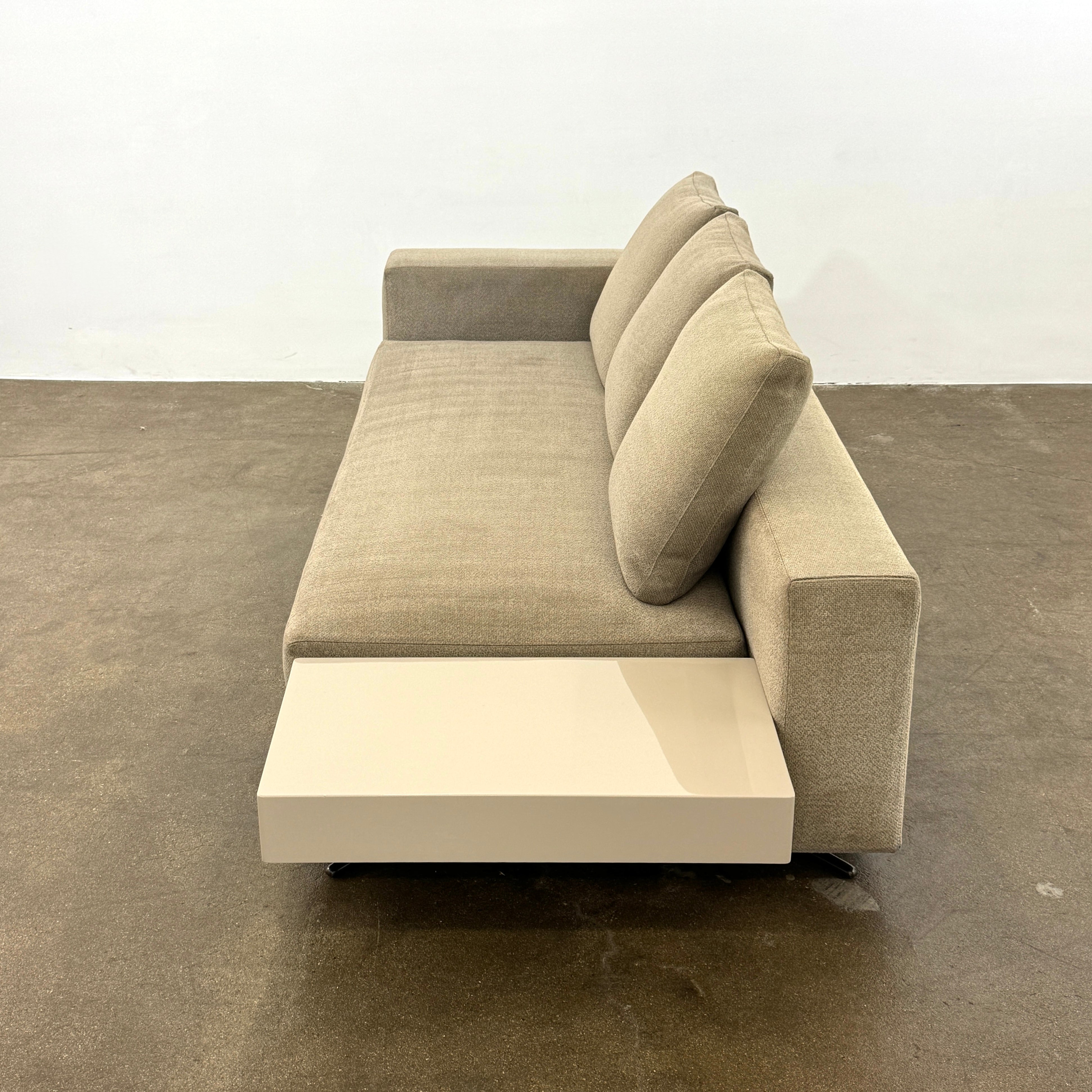 The Minotti White Sofa with Table is a beige three-seater luxury sofa featuring chenille upholstery, wide armrests, three back cushions, and an attached white side table on the right, set against a plain white wall on a brown floor.