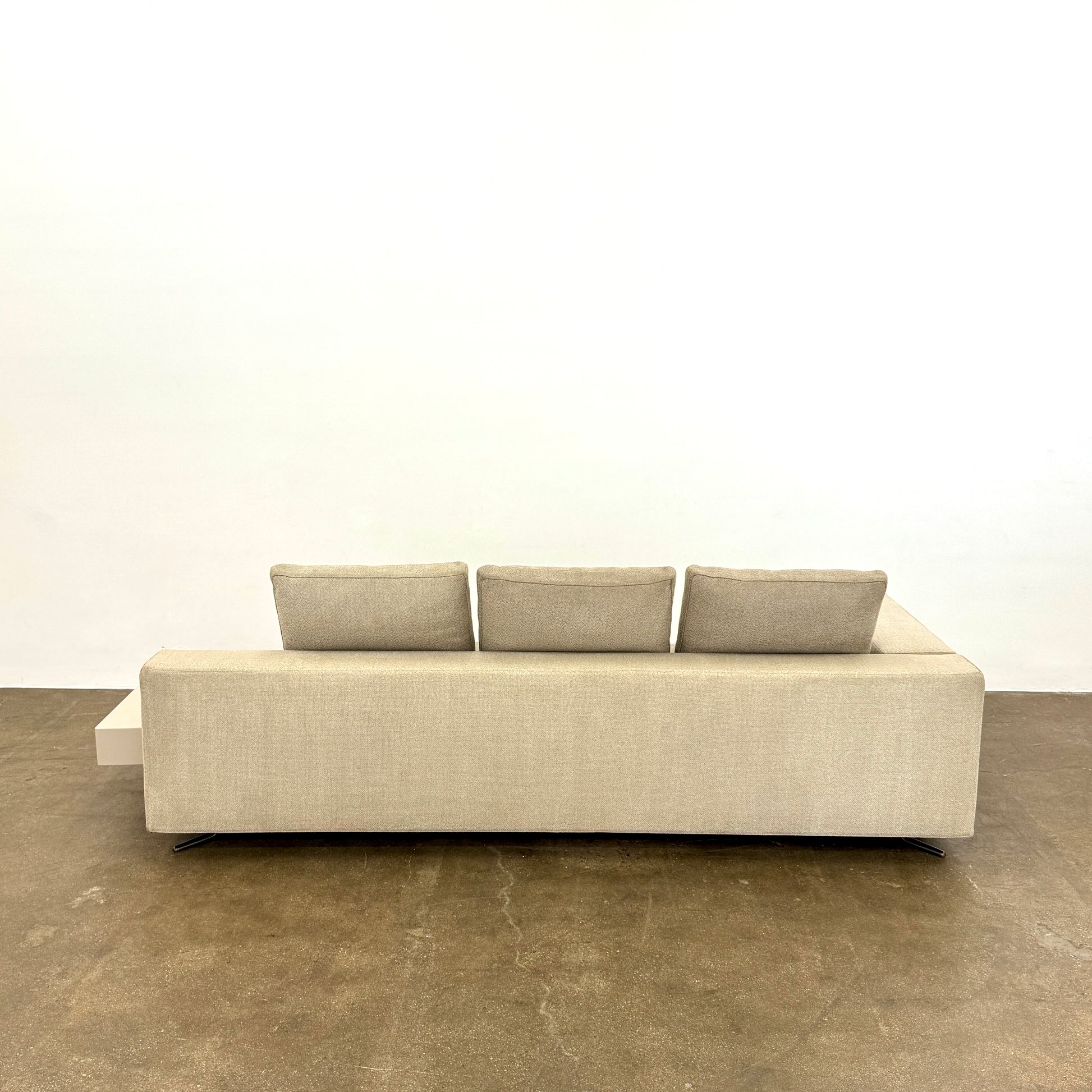 The Minotti White Sofa with Table is a beige three-seater luxury sofa featuring chenille upholstery, wide armrests, three back cushions, and an attached white side table on the right, set against a plain white wall on a brown floor.