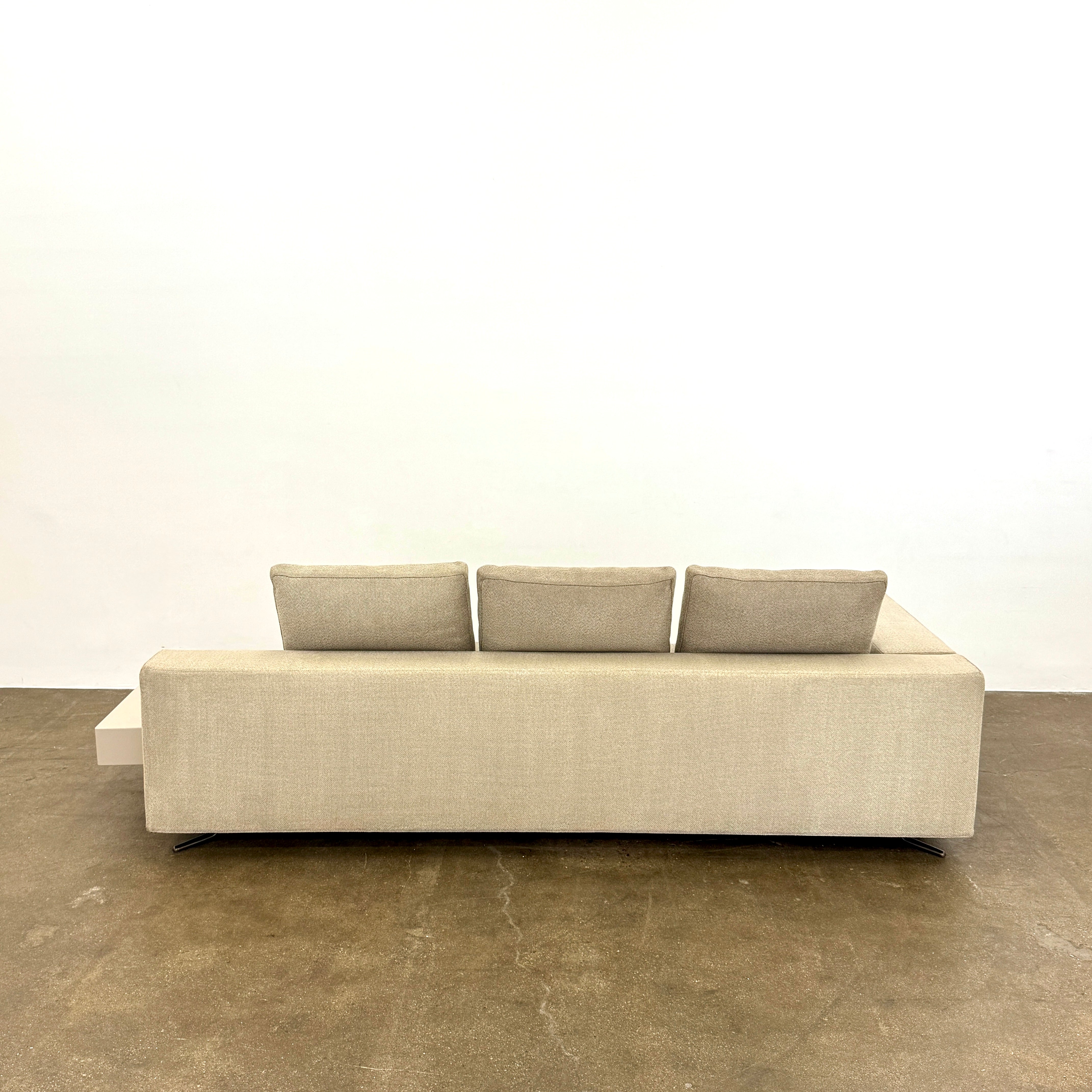 The Minotti White Sofa with Table is a beige three-seater luxury sofa featuring chenille upholstery, wide armrests, three back cushions, and an attached white side table on the right, set against a plain white wall on a brown floor.
