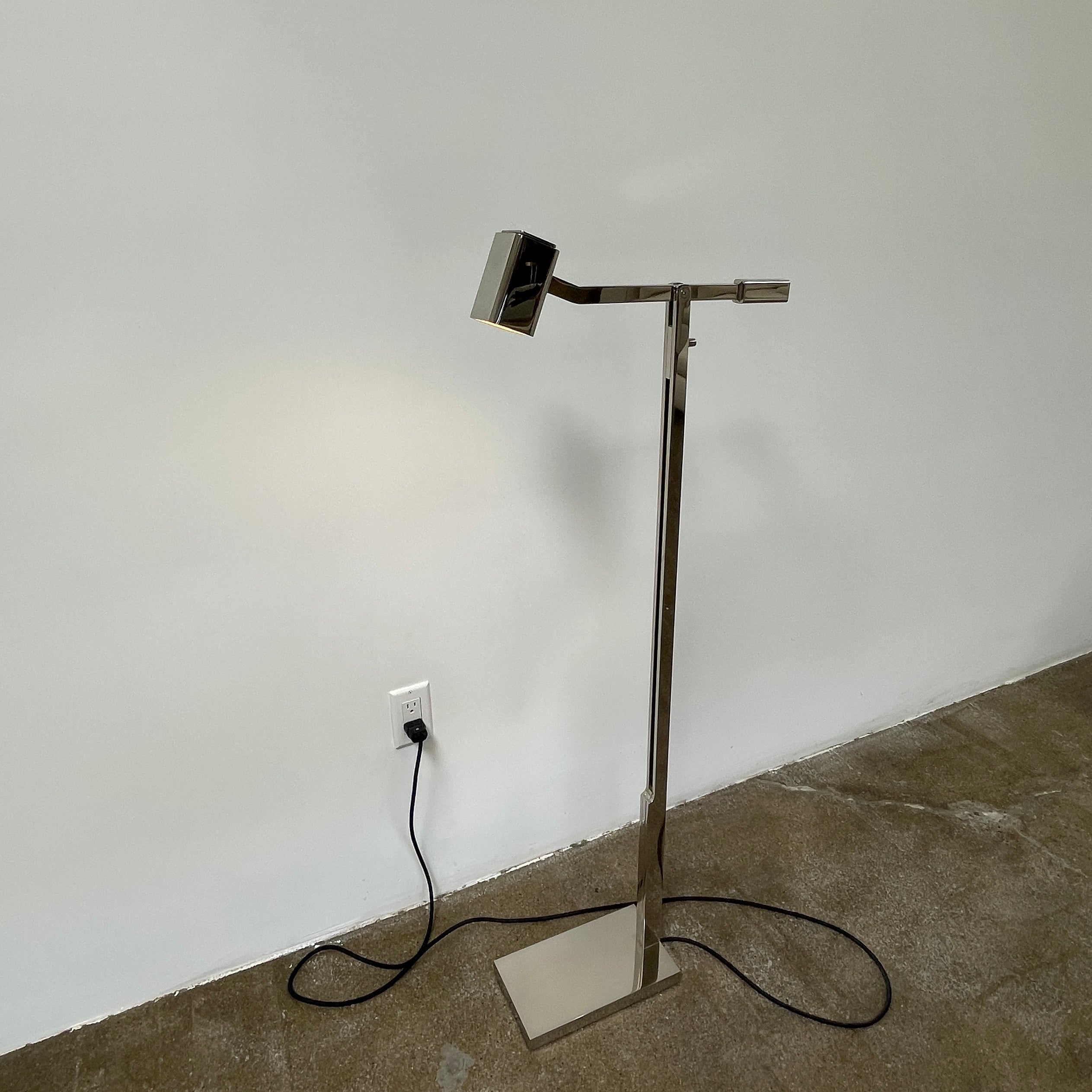 The Holly Hunt Studio Oscar Floor Lamp, by Holly Hunt Studio, features a polished nickel finish with LED lighting, a modern metallic design, an adjustable arm, and a rectangular head. It stands plugged into a wall outlet on a bare concrete floor against a blank wall.