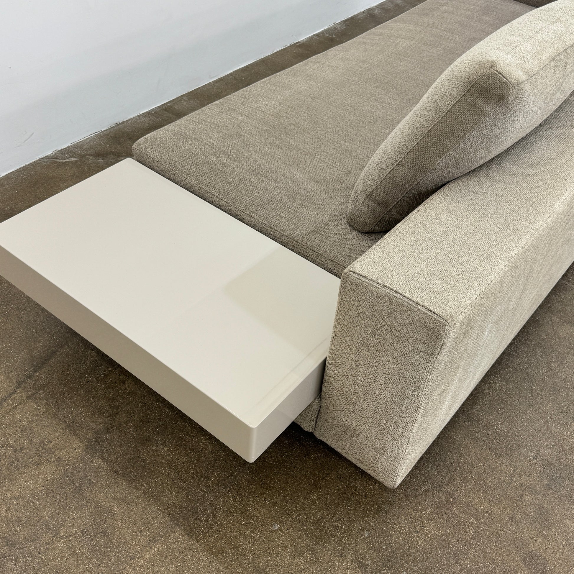 The Minotti White Sofa with Table is a beige three-seater luxury sofa featuring chenille upholstery, wide armrests, three back cushions, and an attached white side table on the right, set against a plain white wall on a brown floor.