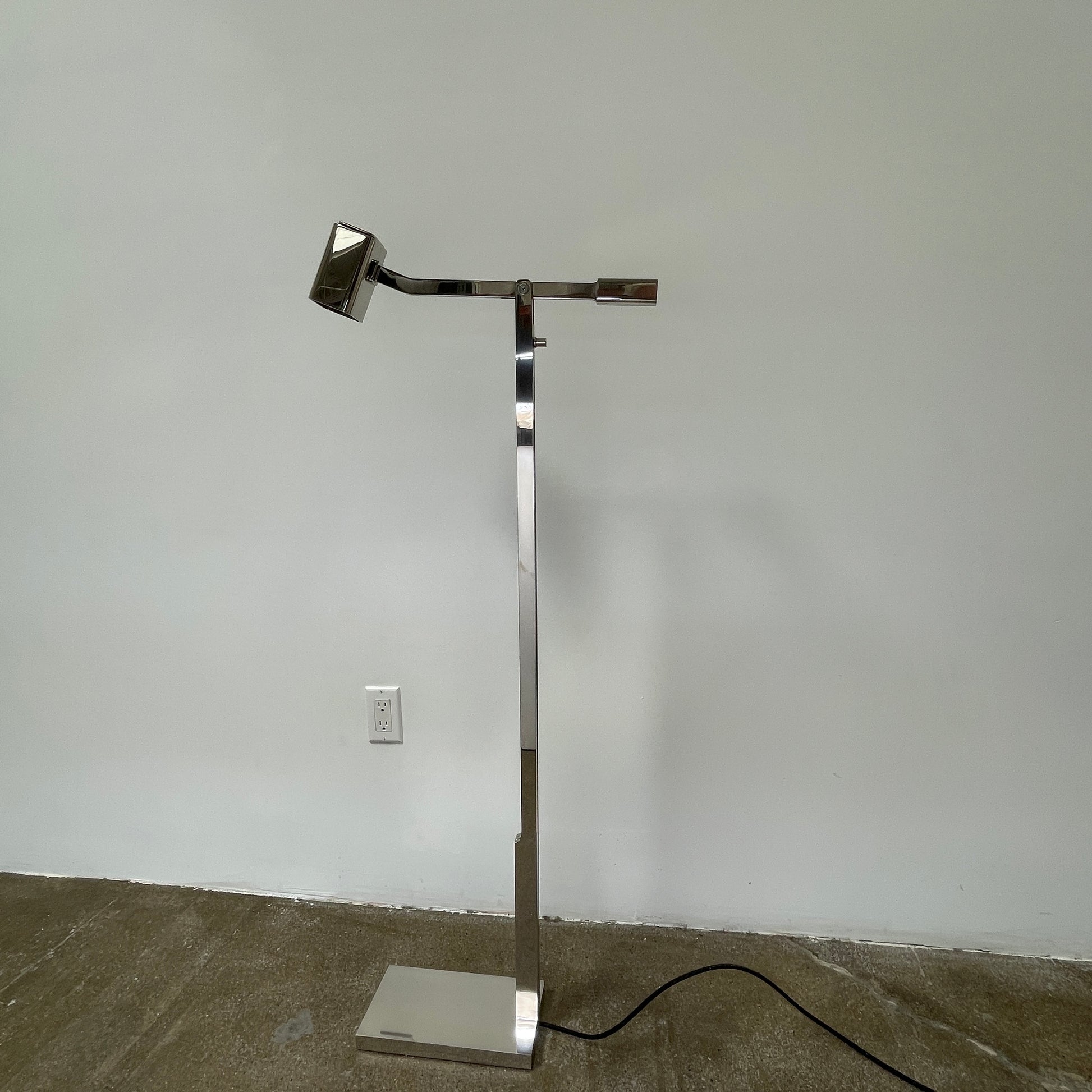 The Holly Hunt Studio Oscar Floor Lamp, by Holly Hunt Studio, features a polished nickel finish with LED lighting, a modern metallic design, an adjustable arm, and a rectangular head. It stands plugged into a wall outlet on a bare concrete floor against a blank wall.