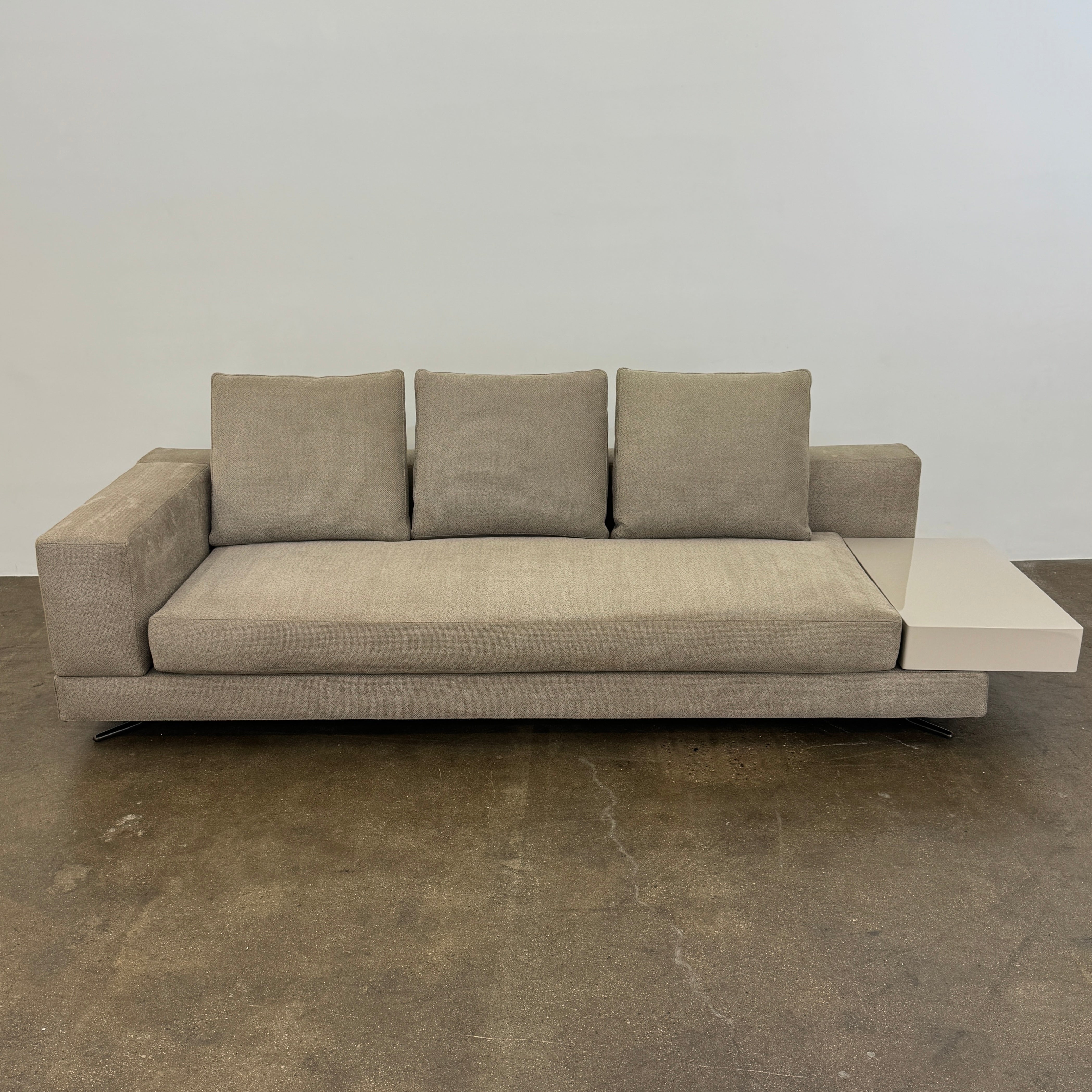 The Minotti White Sofa with Table is a beige three-seater luxury sofa featuring chenille upholstery, wide armrests, three back cushions, and an attached white side table on the right, set against a plain white wall on a brown floor.