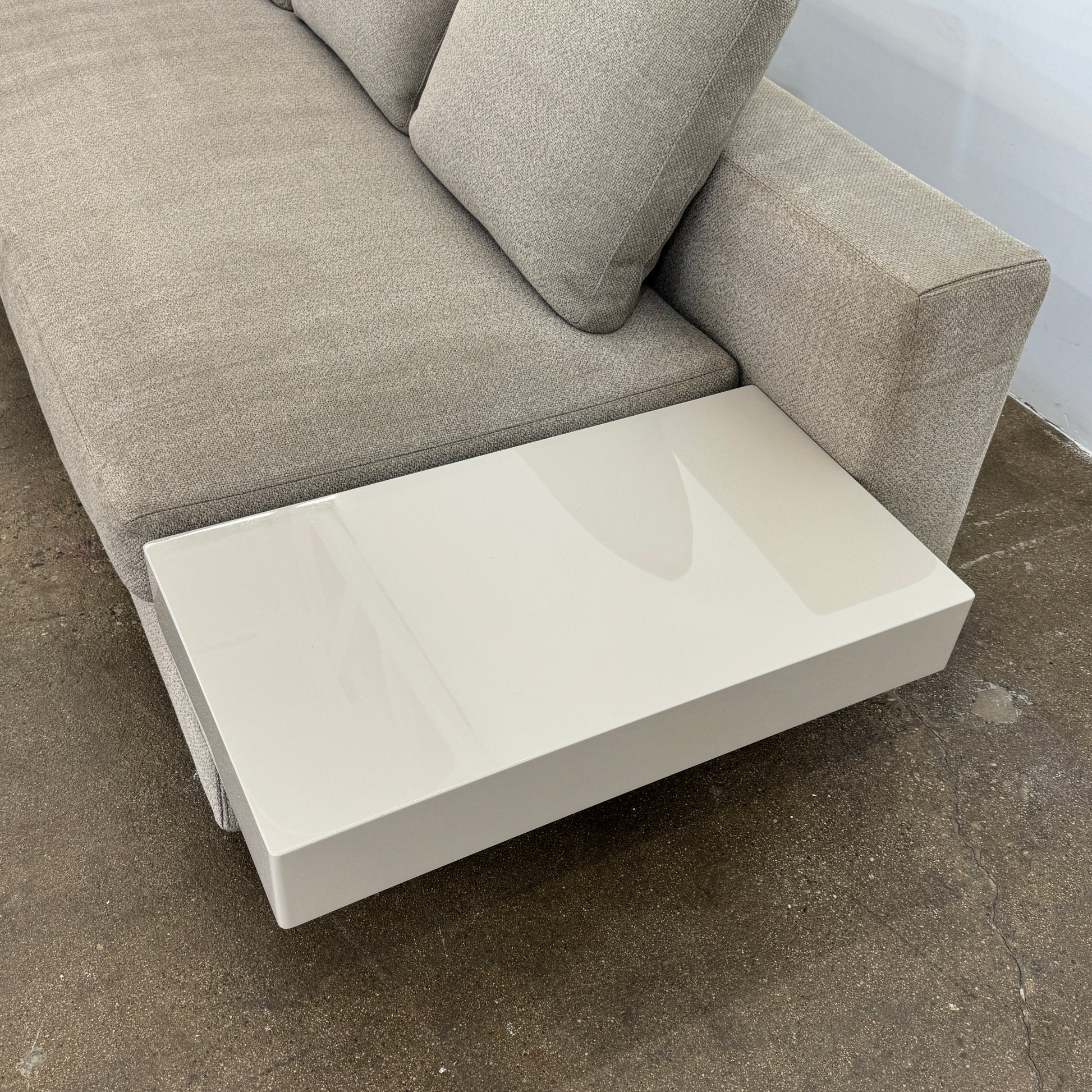 The Minotti White Sofa with Table is a beige three-seater luxury sofa featuring chenille upholstery, wide armrests, three back cushions, and an attached white side table on the right, set against a plain white wall on a brown floor.