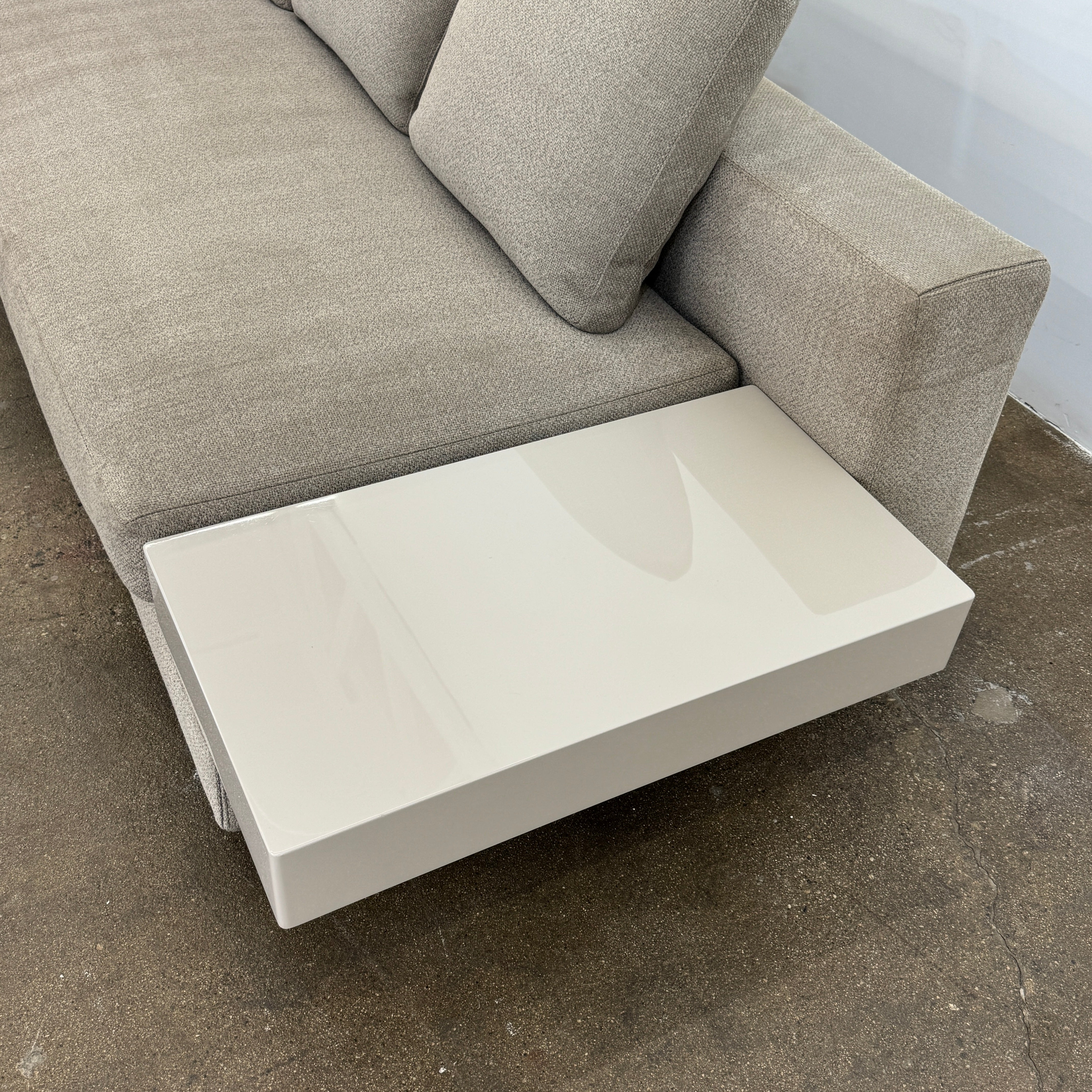 The Minotti White Sofa with Table is a beige three-seater luxury sofa featuring chenille upholstery, wide armrests, three back cushions, and an attached white side table on the right, set against a plain white wall on a brown floor.