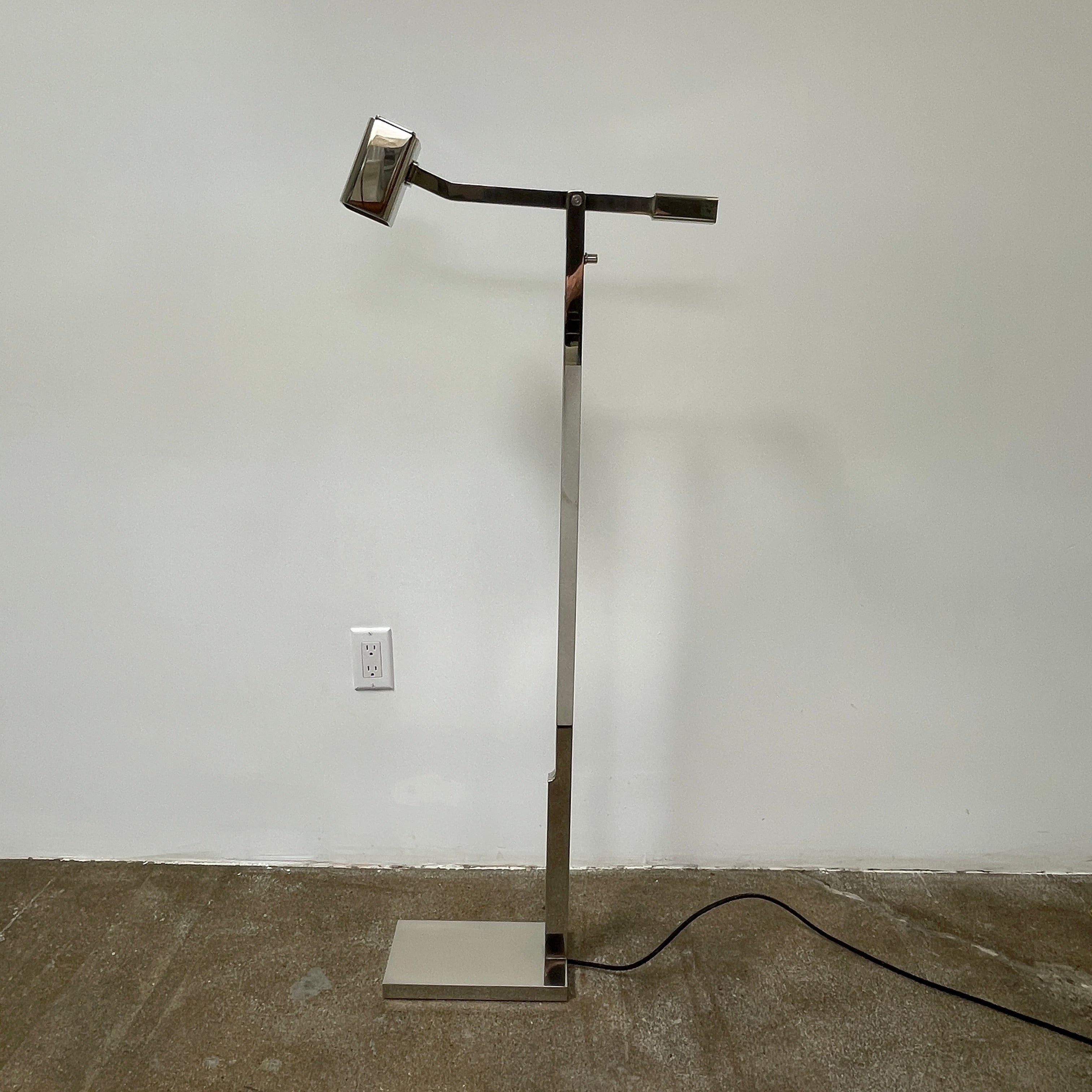 The Holly Hunt Studio Oscar Floor Lamp, by Holly Hunt Studio, features a polished nickel finish with LED lighting, a modern metallic design, an adjustable arm, and a rectangular head. It stands plugged into a wall outlet on a bare concrete floor against a blank wall.