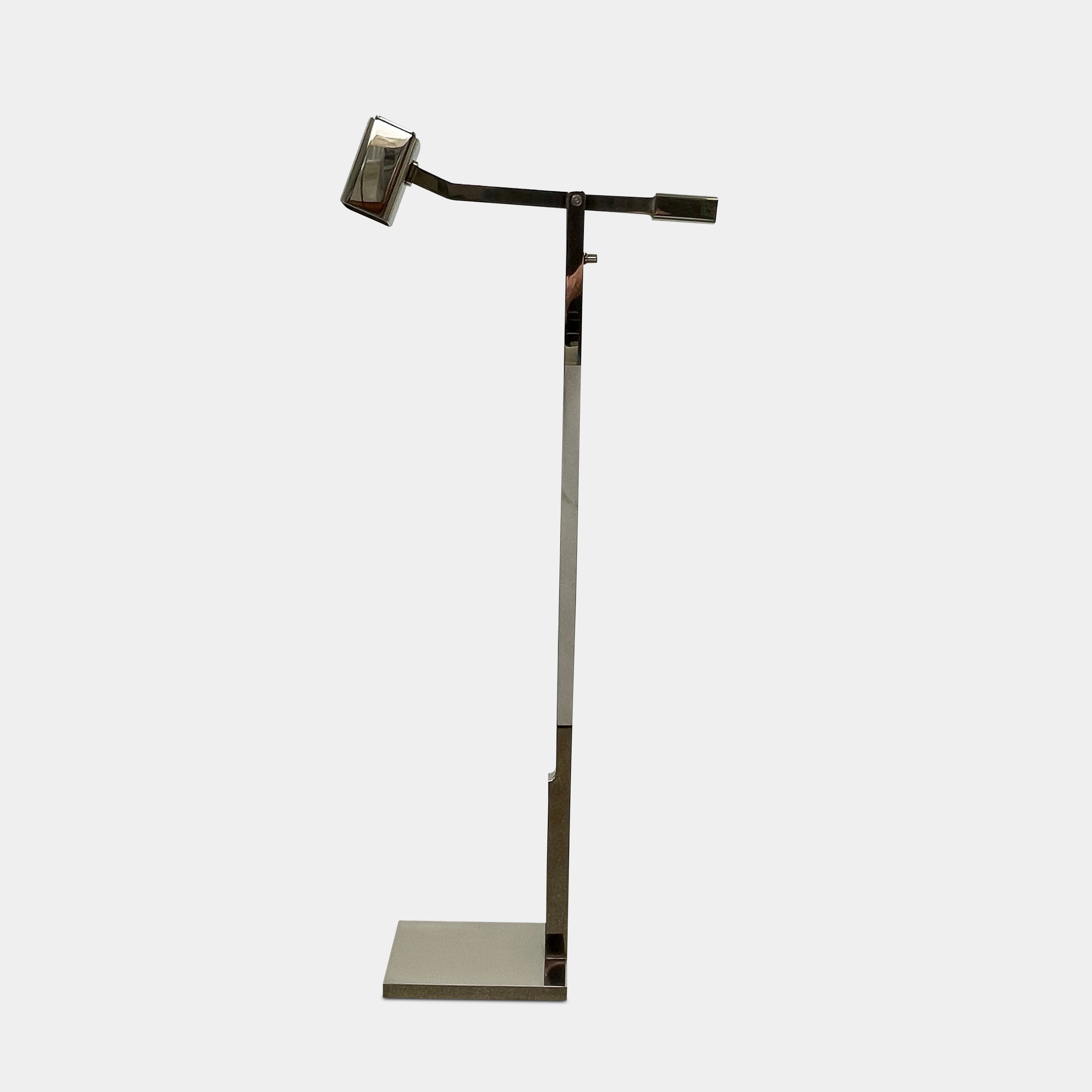 The Holly Hunt Studio Oscar Floor Lamp, by Holly Hunt Studio, features a polished nickel finish with LED lighting, a modern metallic design, an adjustable arm, and a rectangular head. It stands plugged into a wall outlet on a bare concrete floor against a blank wall.