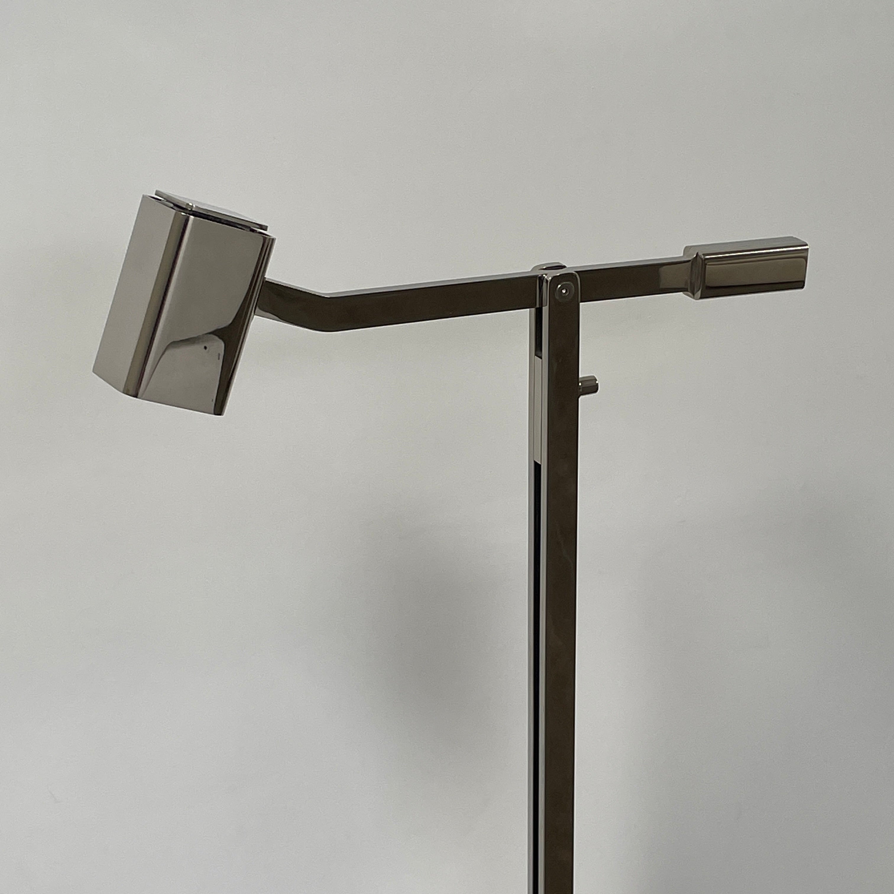 The Holly Hunt Studio Oscar Floor Lamp, by Holly Hunt Studio, features a polished nickel finish with LED lighting, a modern metallic design, an adjustable arm, and a rectangular head. It stands plugged into a wall outlet on a bare concrete floor against a blank wall.