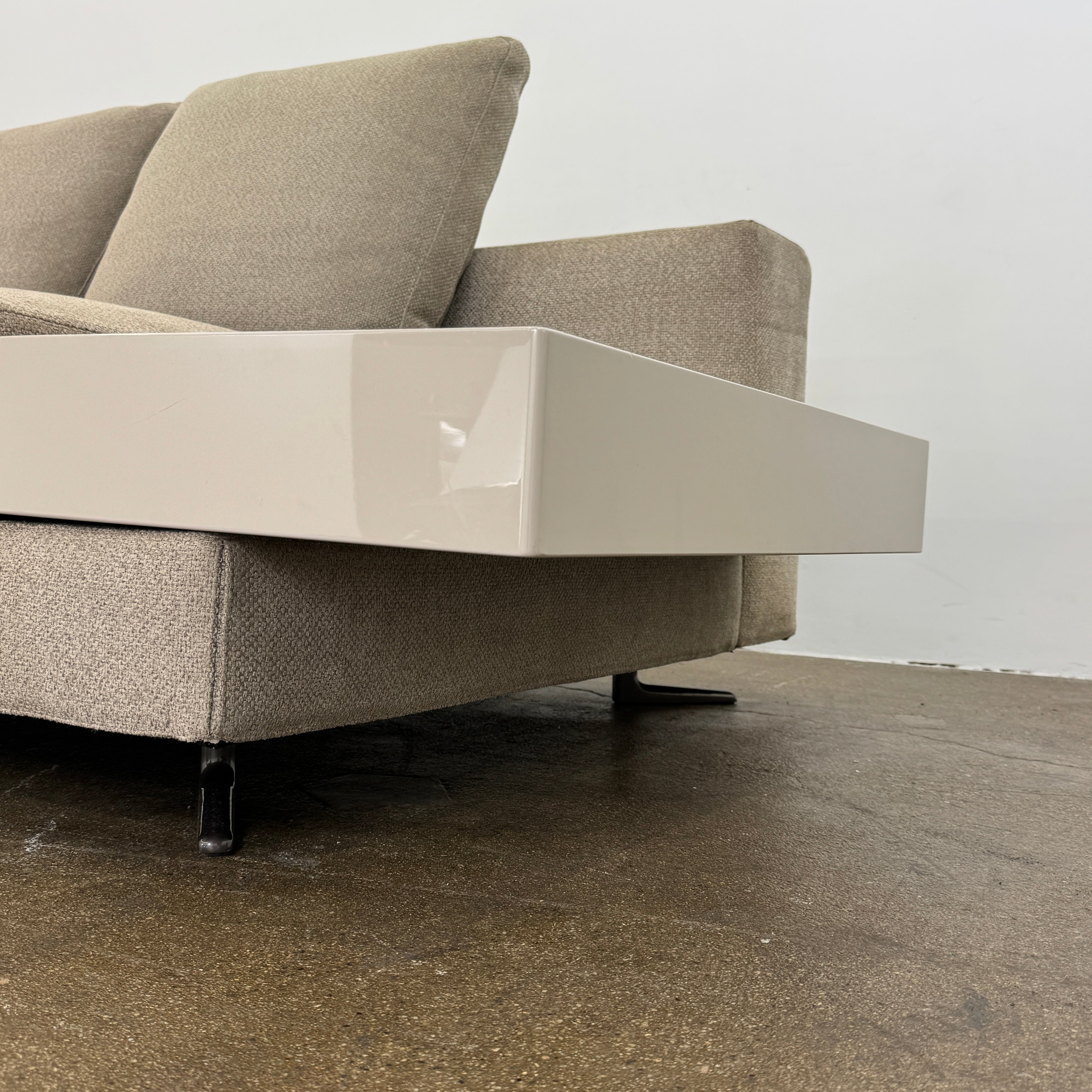 The Minotti White Sofa with Table is a beige three-seater luxury sofa featuring chenille upholstery, wide armrests, three back cushions, and an attached white side table on the right, set against a plain white wall on a brown floor.