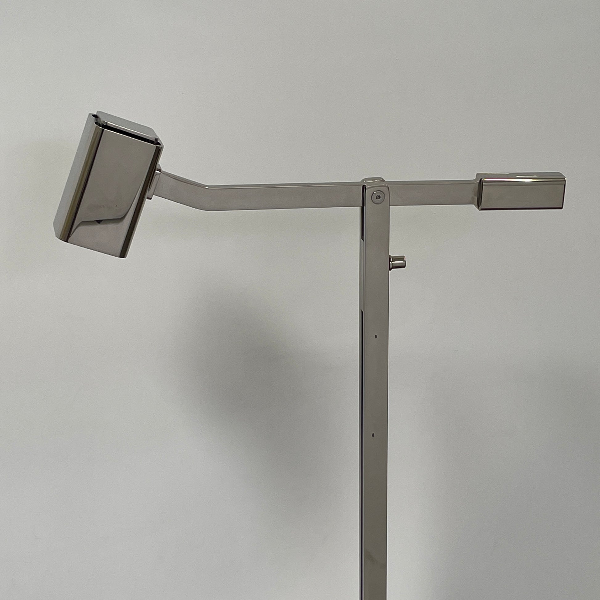 The Holly Hunt Studio Oscar Floor Lamp, by Holly Hunt Studio, features a polished nickel finish with LED lighting, a modern metallic design, an adjustable arm, and a rectangular head. It stands plugged into a wall outlet on a bare concrete floor against a blank wall.