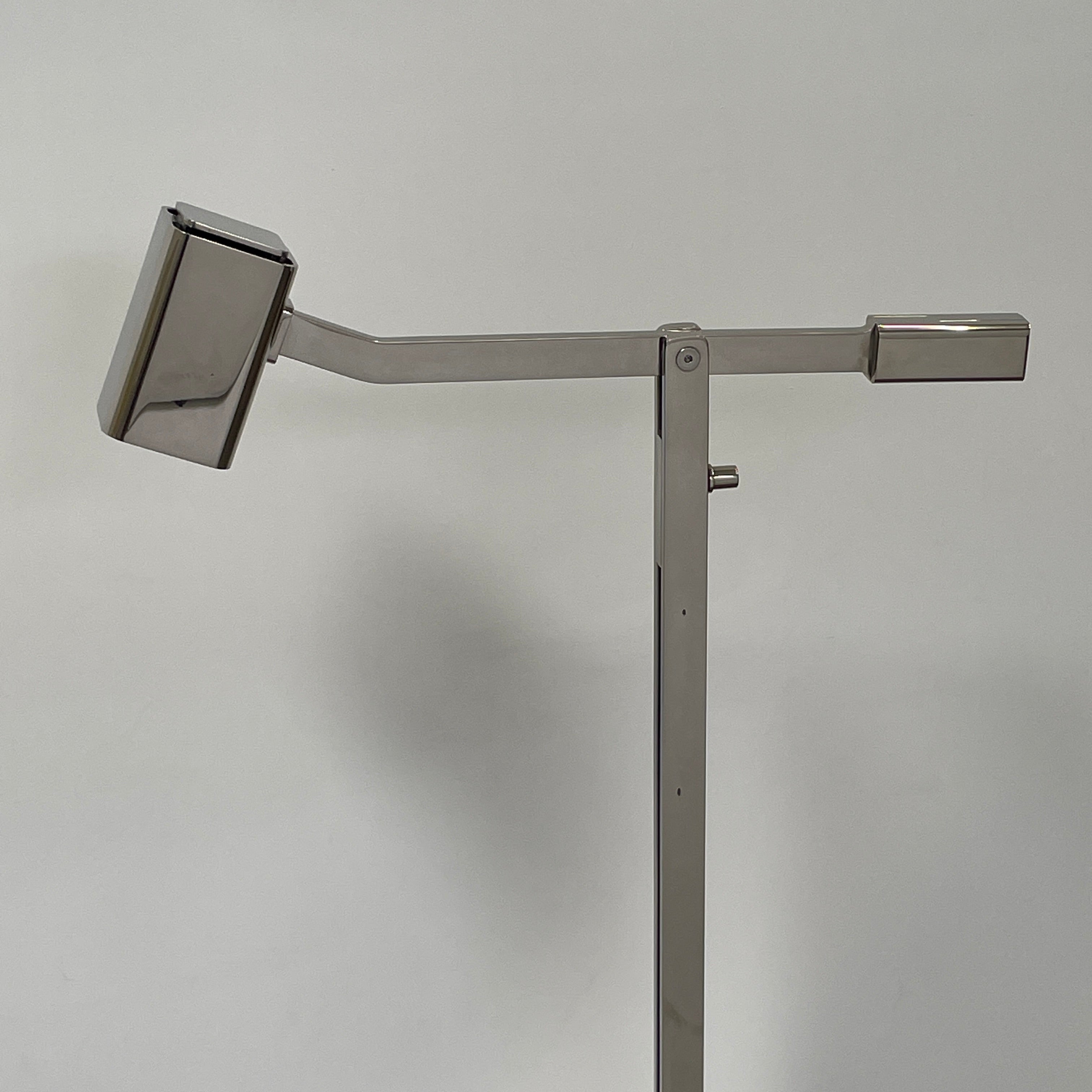 The Holly Hunt Studio Oscar Floor Lamp, by Holly Hunt Studio, features a polished nickel finish with LED lighting, a modern metallic design, an adjustable arm, and a rectangular head. It stands plugged into a wall outlet on a bare concrete floor against a blank wall.