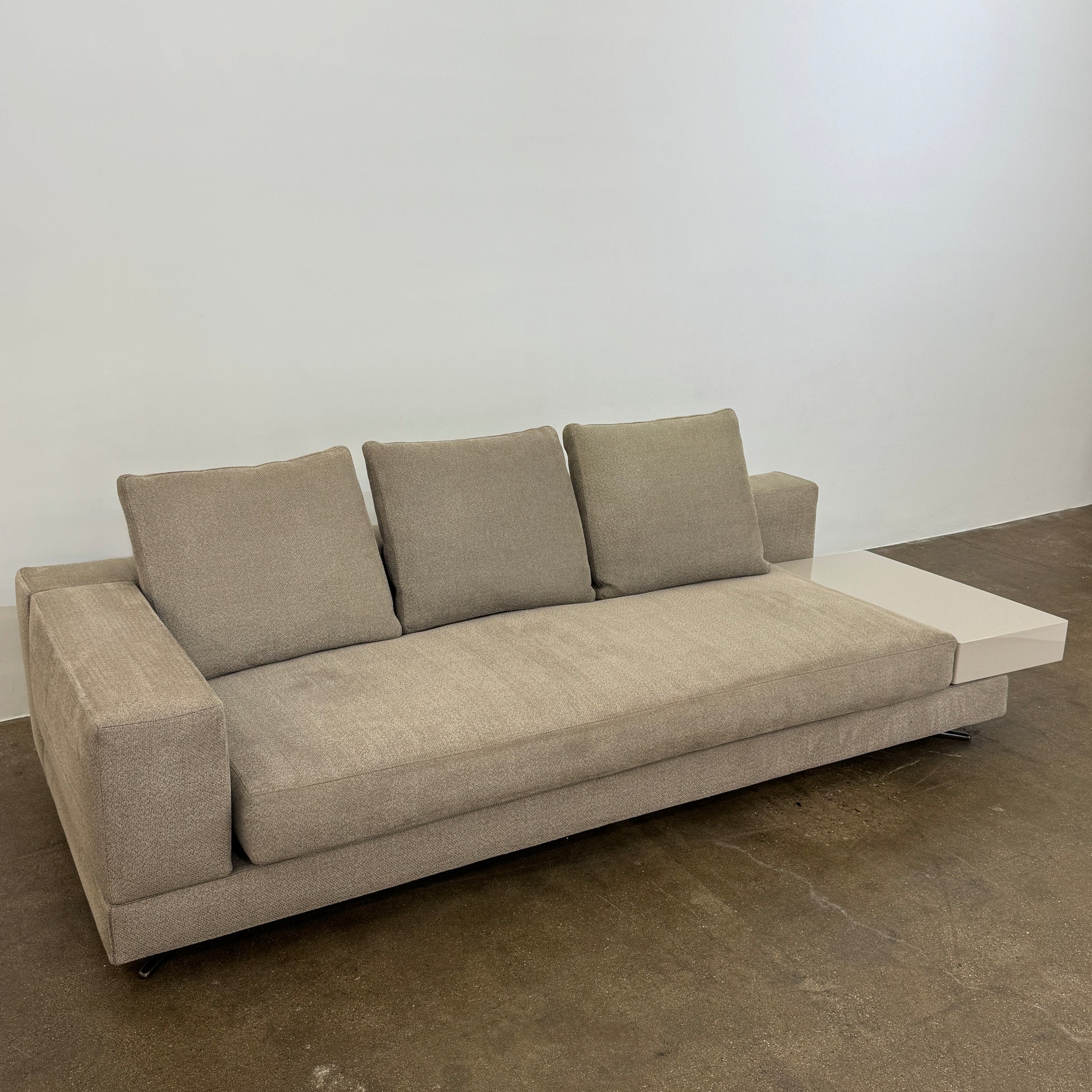 The Minotti White Sofa with Table is a beige three-seater luxury sofa featuring chenille upholstery, wide armrests, three back cushions, and an attached white side table on the right, set against a plain white wall on a brown floor.