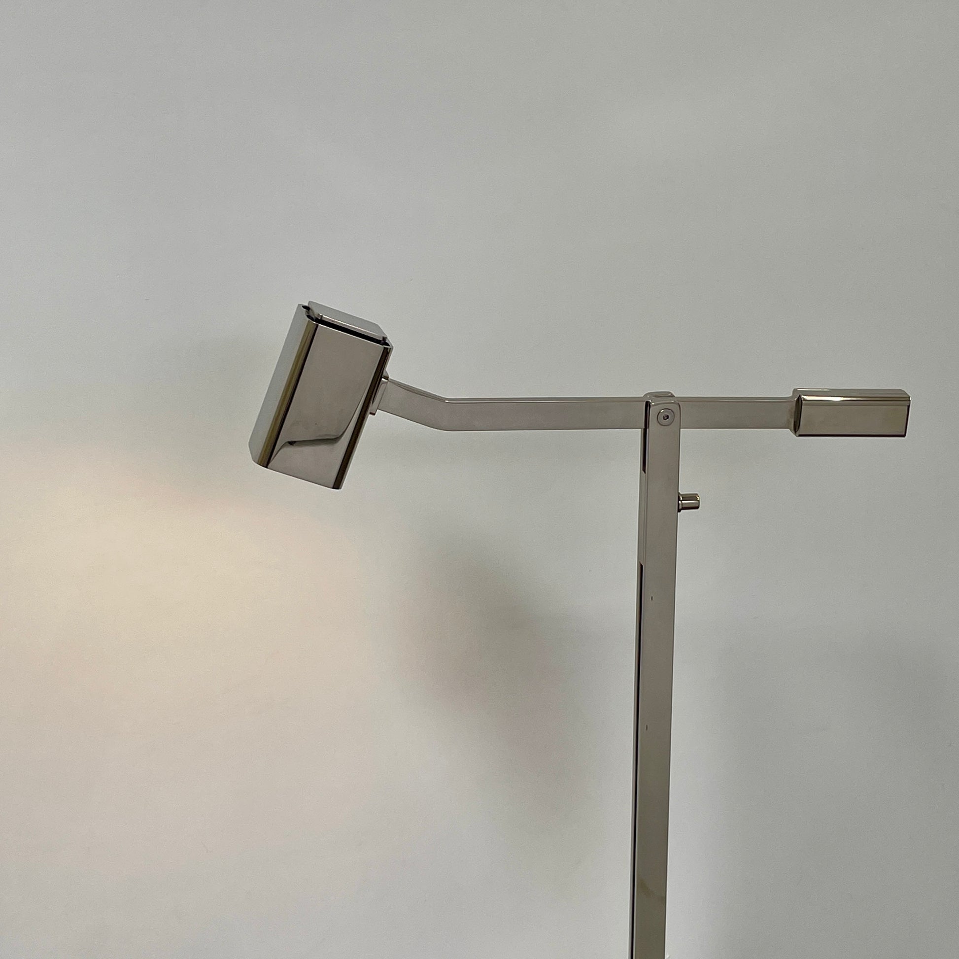 The Holly Hunt Studio Oscar Floor Lamp, by Holly Hunt Studio, features a polished nickel finish with LED lighting, a modern metallic design, an adjustable arm, and a rectangular head. It stands plugged into a wall outlet on a bare concrete floor against a blank wall.