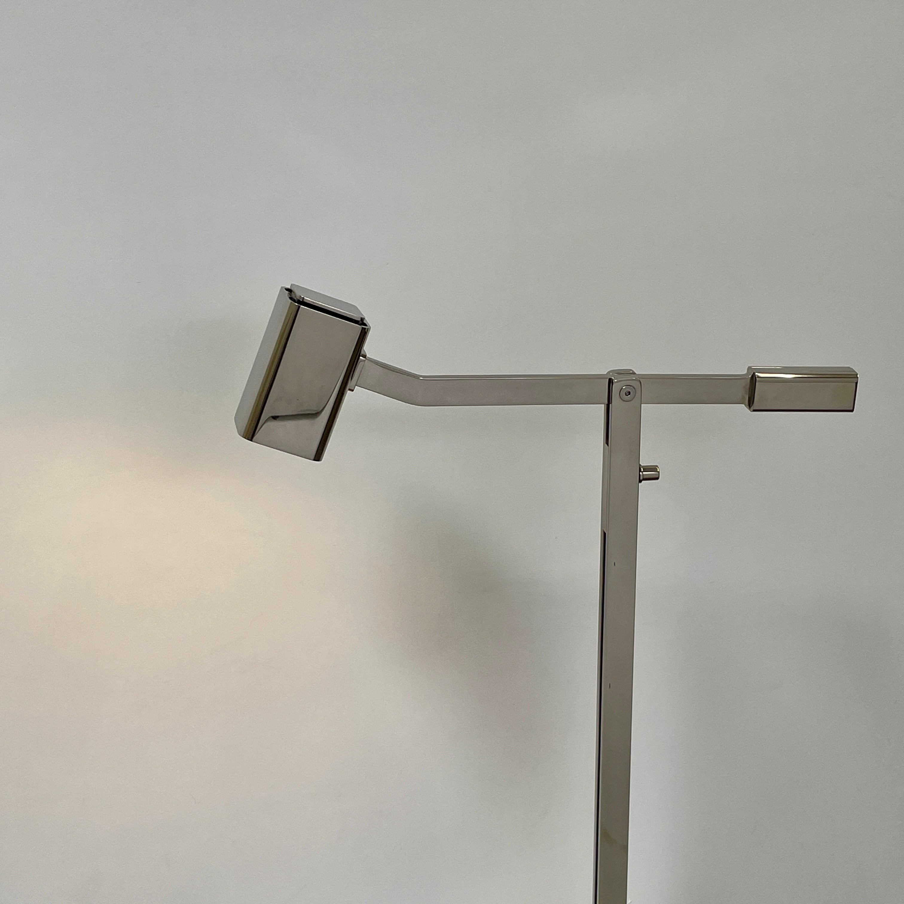 The Holly Hunt Studio Oscar Floor Lamp, by Holly Hunt Studio, features a polished nickel finish with LED lighting, a modern metallic design, an adjustable arm, and a rectangular head. It stands plugged into a wall outlet on a bare concrete floor against a blank wall.