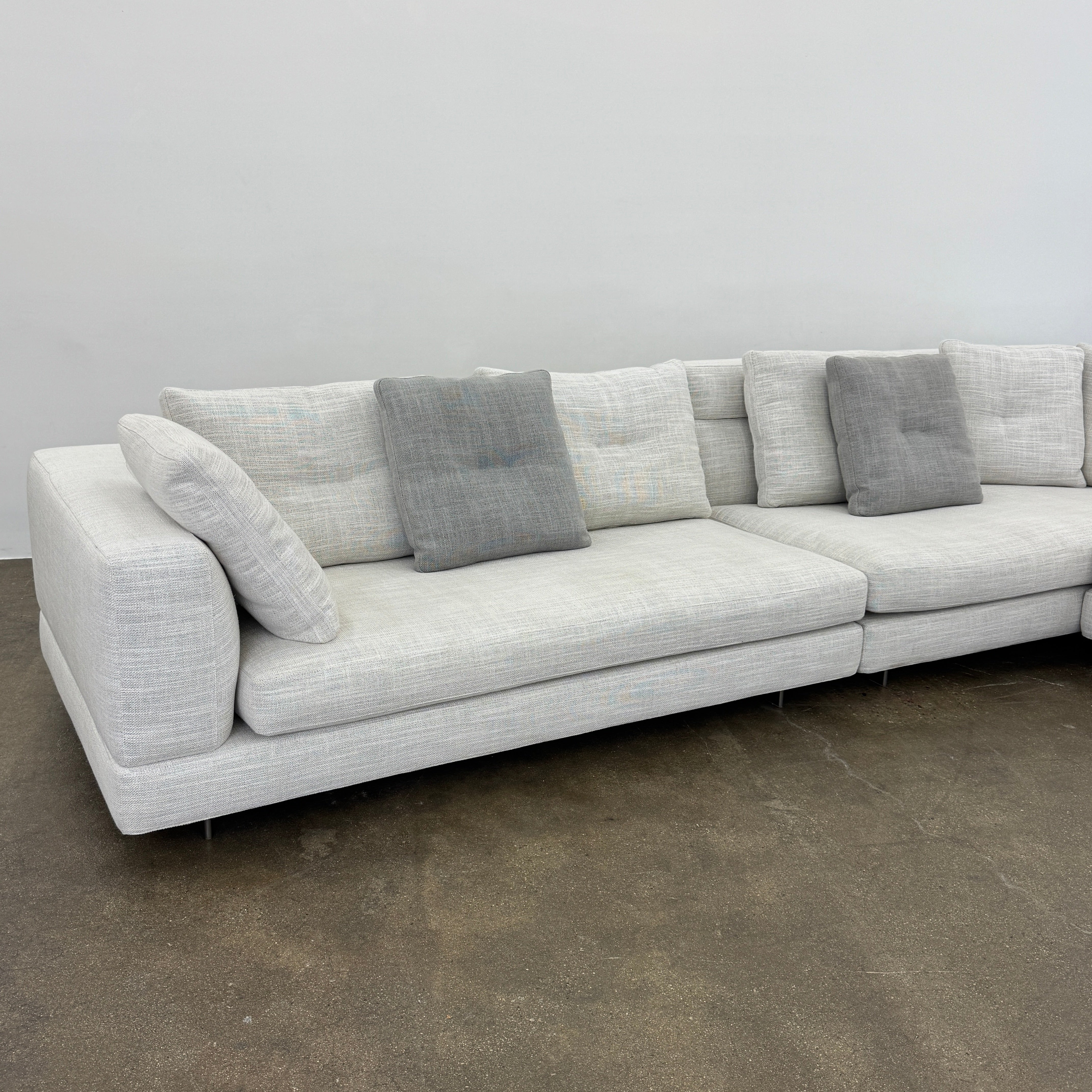 The Minotti Alexander 3 Piece Sectional by Minotti features an L-shaped design in light beige linen blend upholstery with multiple gray and beige throw pillows, set against a plain white background.