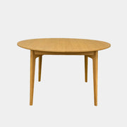 A Case Dulwich Extension Table, round and wooden with four legs, stands on a concrete floor against a plain white wall.