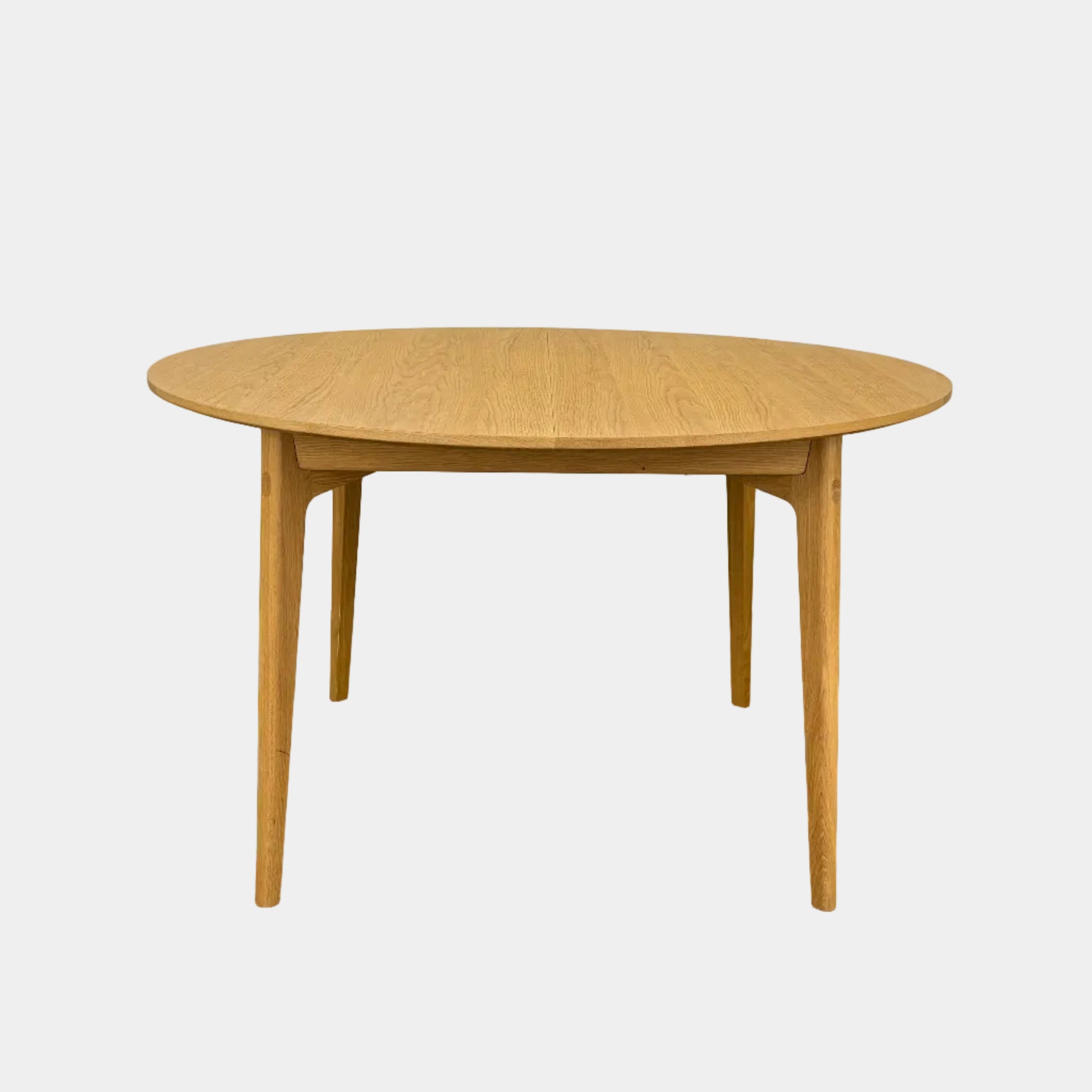 A Case Dulwich Extension Table, round and wooden with four legs, stands on a concrete floor against a plain white wall.