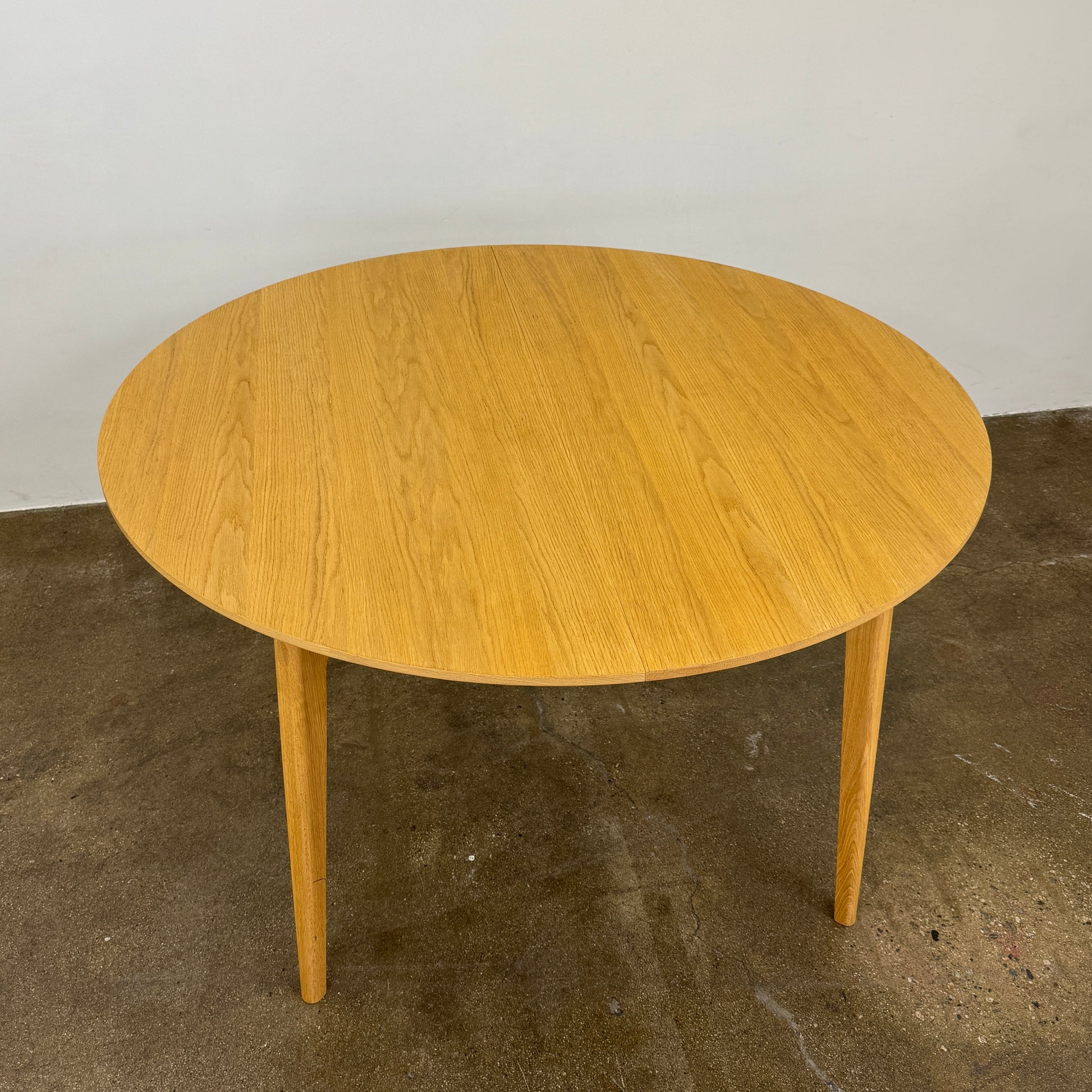 A Case Dulwich Extension Table, round and wooden with four legs, stands on a concrete floor against a plain white wall.