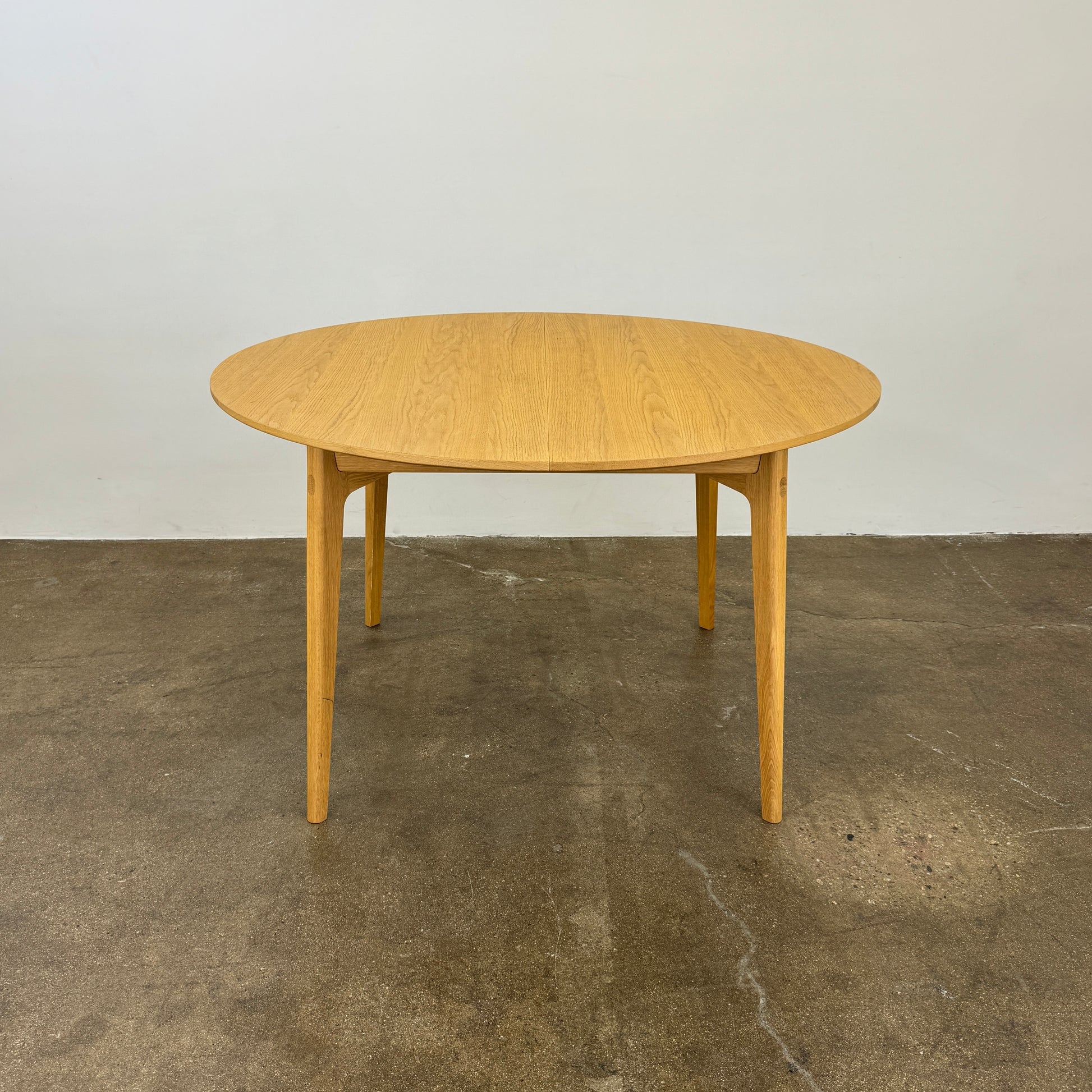 A Case Dulwich Extension Table, round and wooden with four legs, stands on a concrete floor against a plain white wall.