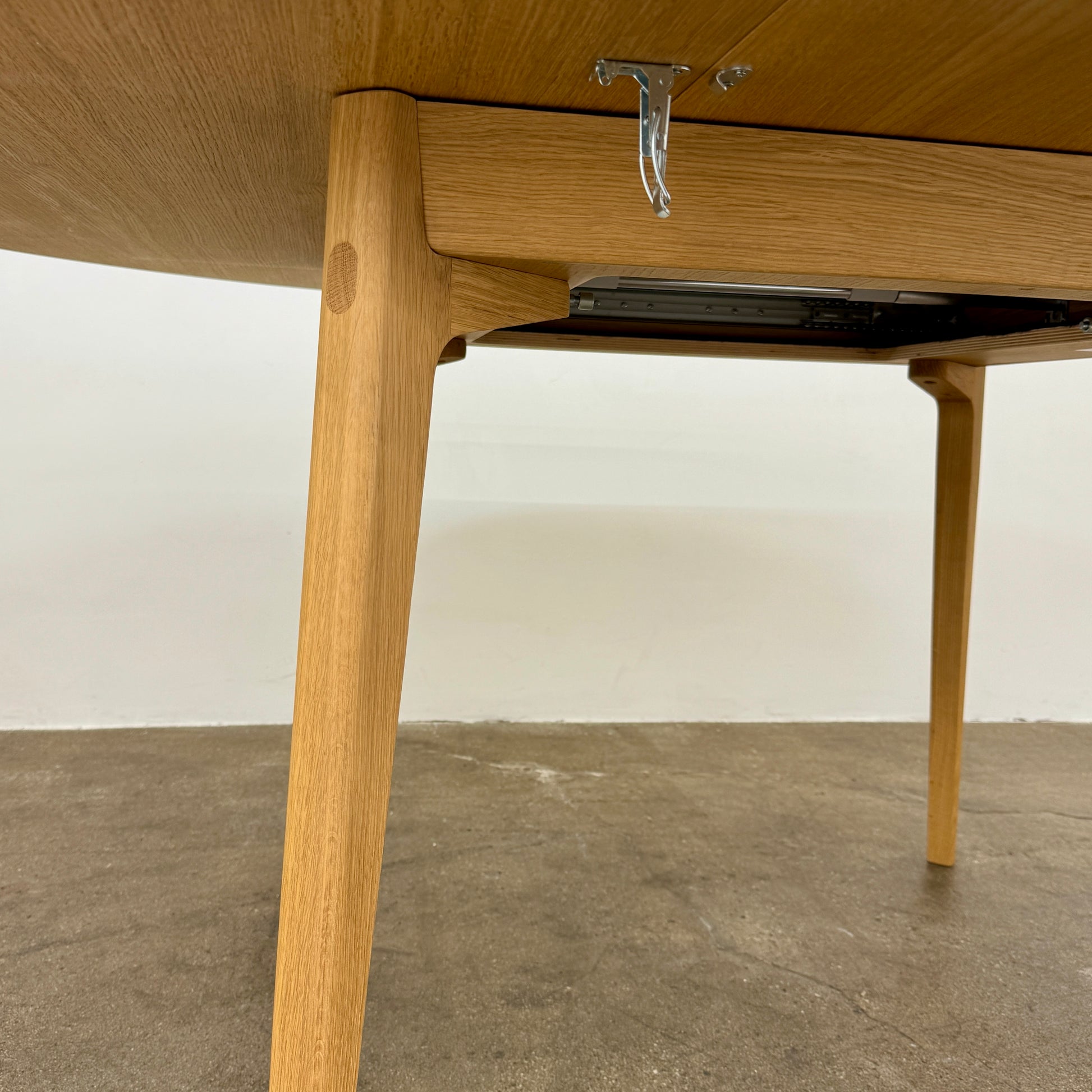 A Case Dulwich Extension Table, round and wooden with four legs, stands on a concrete floor against a plain white wall.