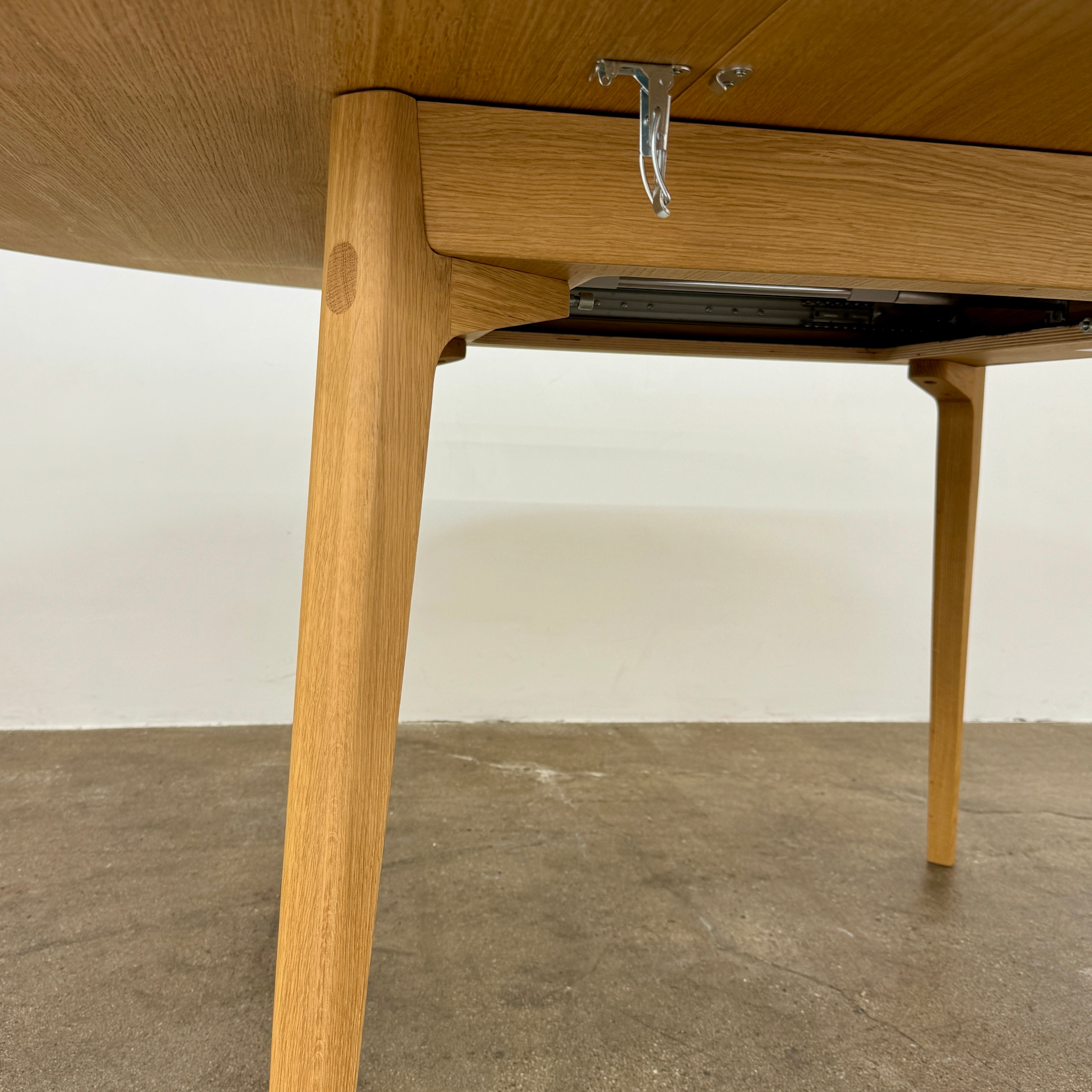 A Case Dulwich Extension Table, round and wooden with four legs, stands on a concrete floor against a plain white wall.